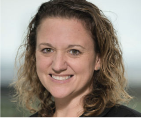 A woman with curly brown hair smiling in front of a blurred outdoor background.