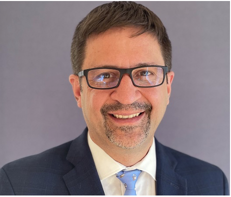 A smiling man wearing glasses, a dark suit, white shirt, and light blue tie, against a gray background.