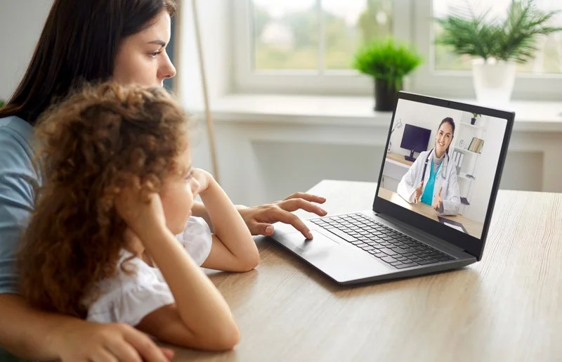 A woman and a young girl are watching a doctor on a laptop during a video call in a bright room with a window and potted plants.