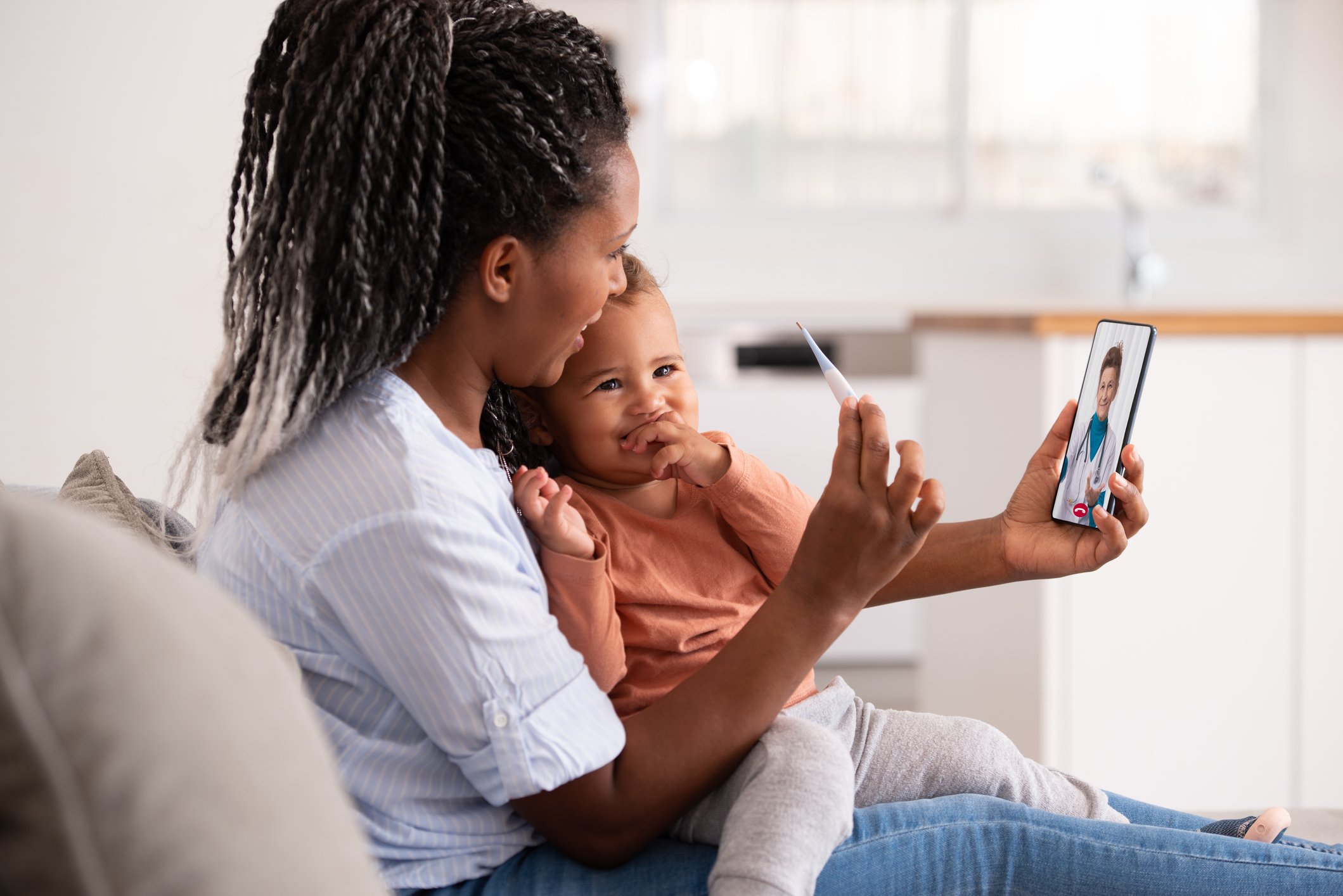 A woman and a young girl are sitting on a couch during a video call with a doctor on a smartphone. The woman is smiling and holding a thermometer, while the girl is hugging her and looking at the phone.