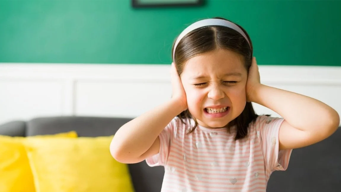 A young girl covering her ears and grimacing, possibly upset or distressed, sitting on a sofa with yellow cushions in the background.