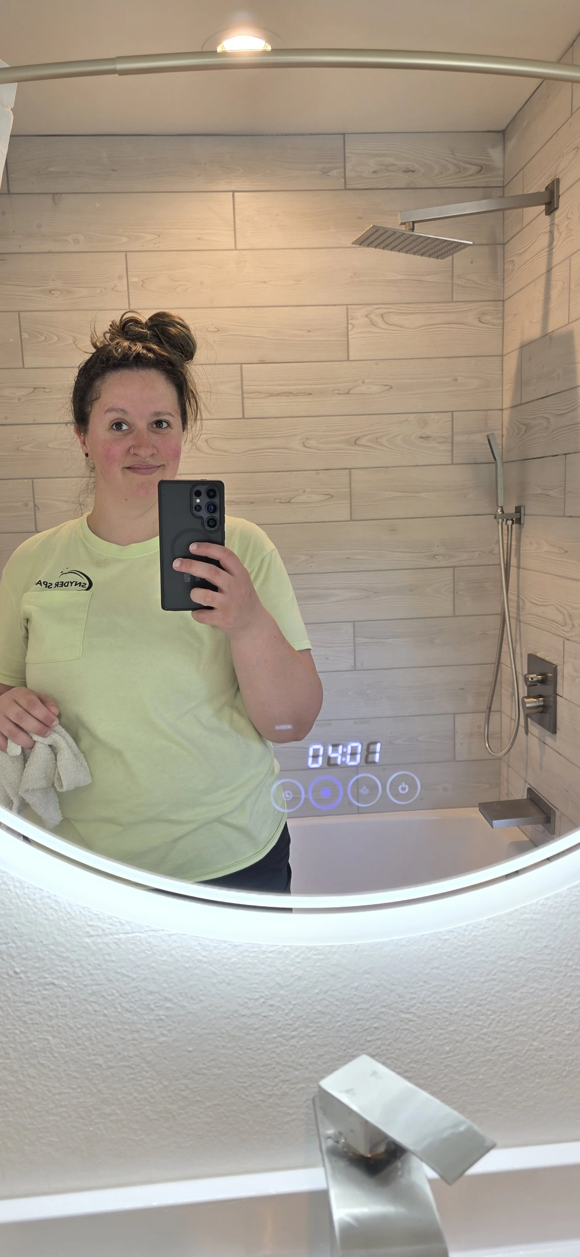 A woman taking a mirror selfie in a bathroom, wearing a yellow t-shirt and holding a towel, with a wooden wall, shower, and digital clock display in the background.