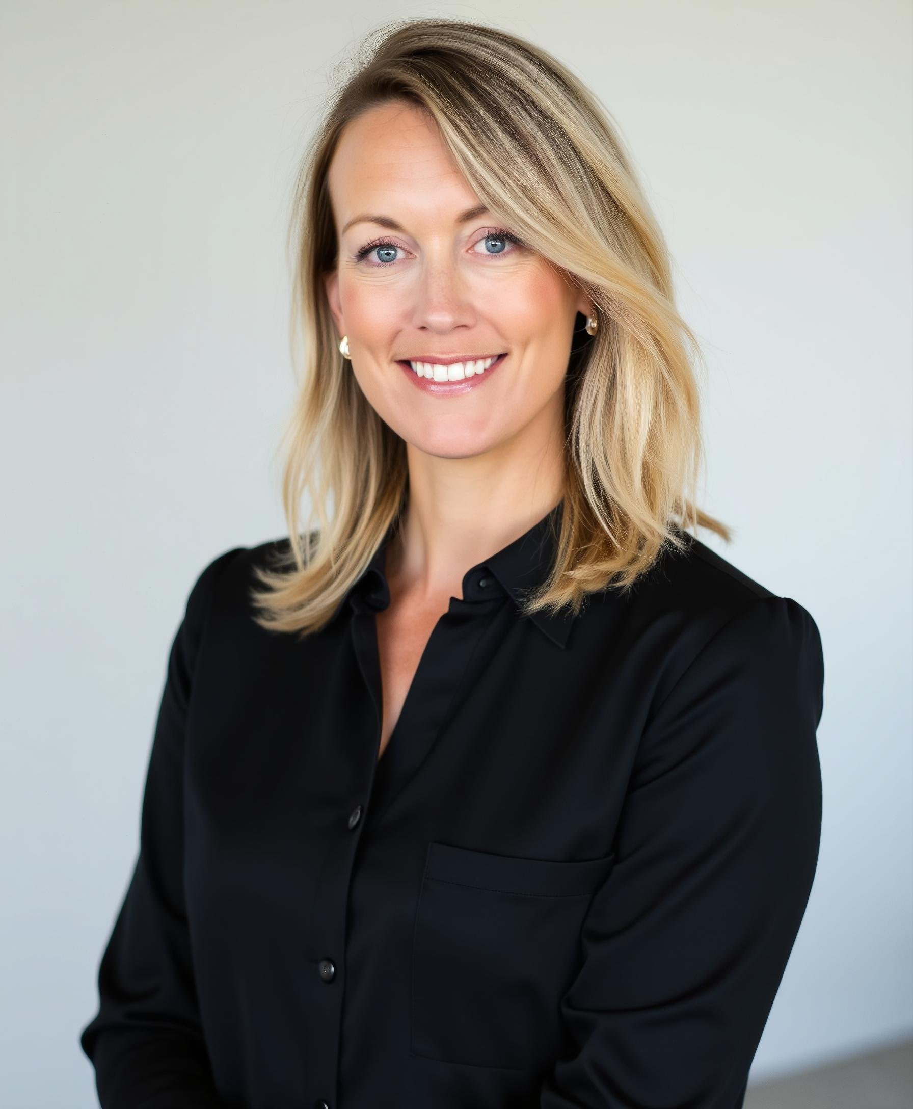 A smiling woman with shoulder-length blonde hair, wearing a black blouse, standing against a plain background.