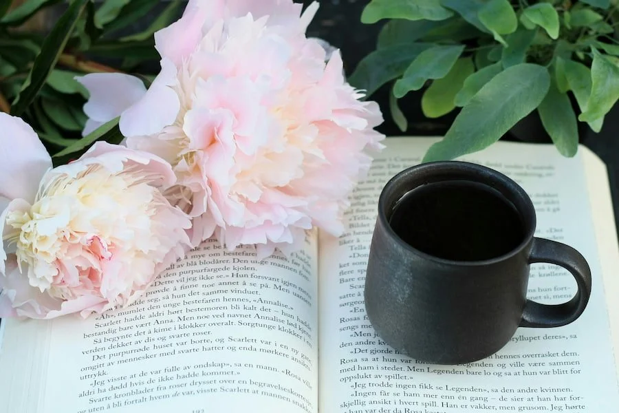 An open book resting beneath soft pink peonies and a dark ceramic mug — a quiet, pleasurable moment of reading