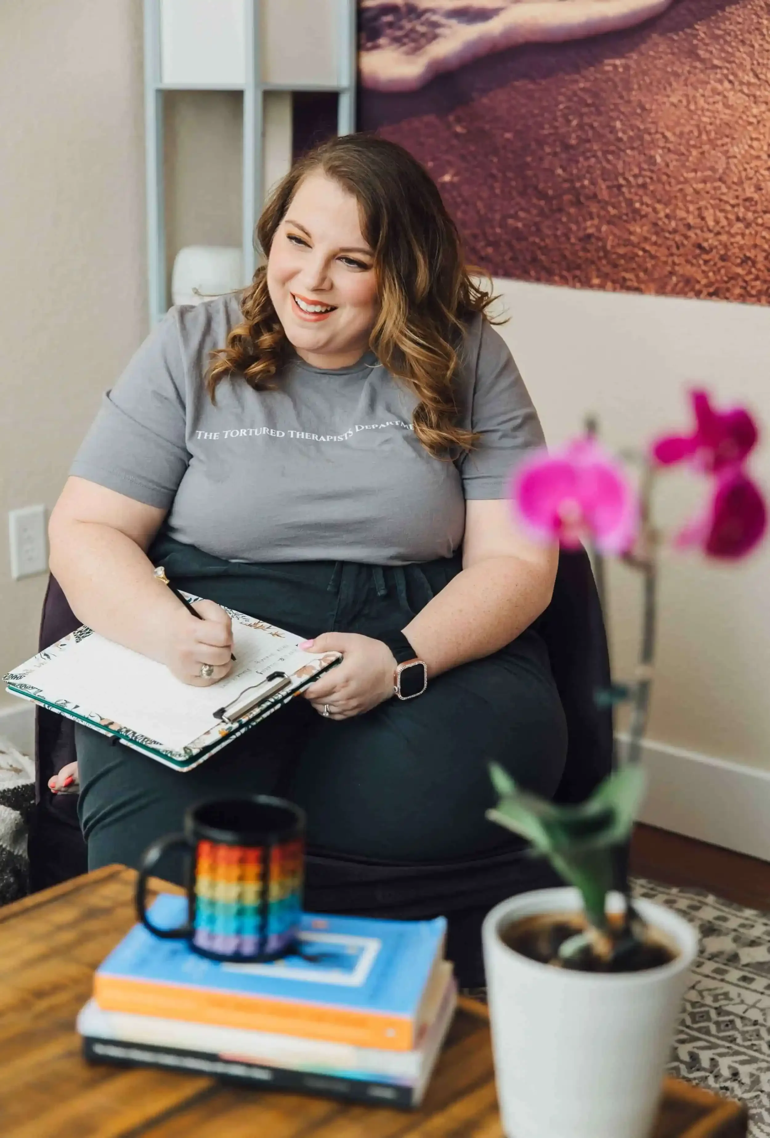 Close-up of Lauren McKay taking notes on a floral notepad with a diamond-topped pen. In the foreground, a wooden coffee table topped with trauma therapy books, a 'Safe Space' mug, creating a warm and welcoming therapy environment.