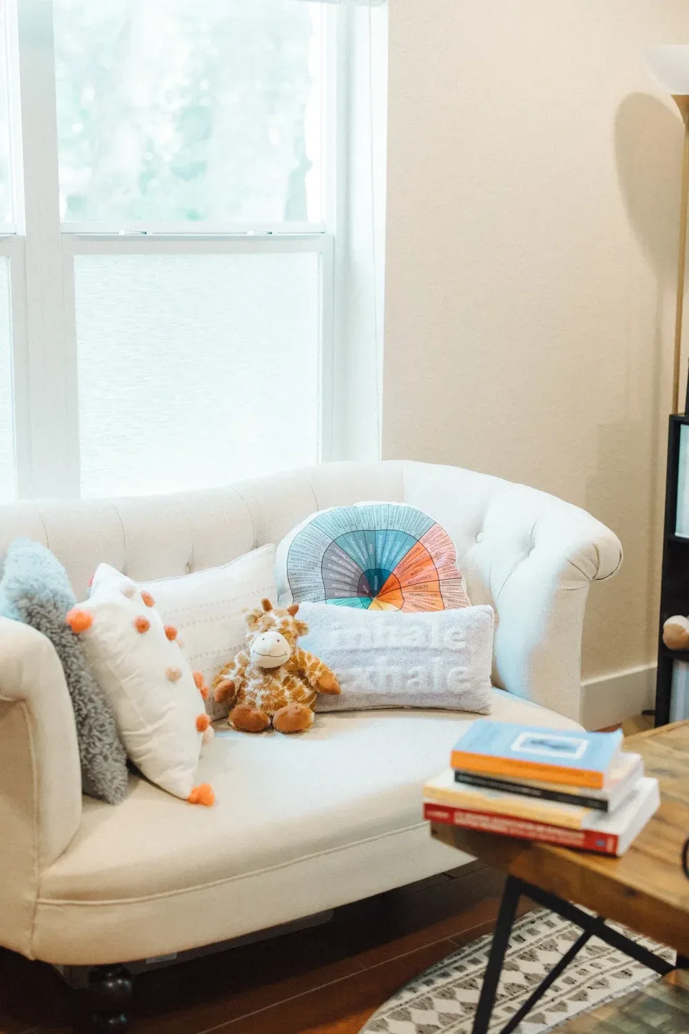 Cozy therapy office corner with a cream tufted loveseat, a colorful emotion wheel pillow, plush giraffe toy, and 'inhale exhale' cushion beneath a large window. In the foreground, atop a wooden coffee table sit trauma therapy books.