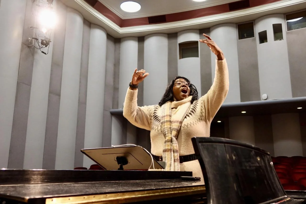 A woman in a beige sweater and scarf is conducting or leading a musical rehearsal in a concert hall, standing behind a music stand and a piano, with her arms raised, expressive and joyful.