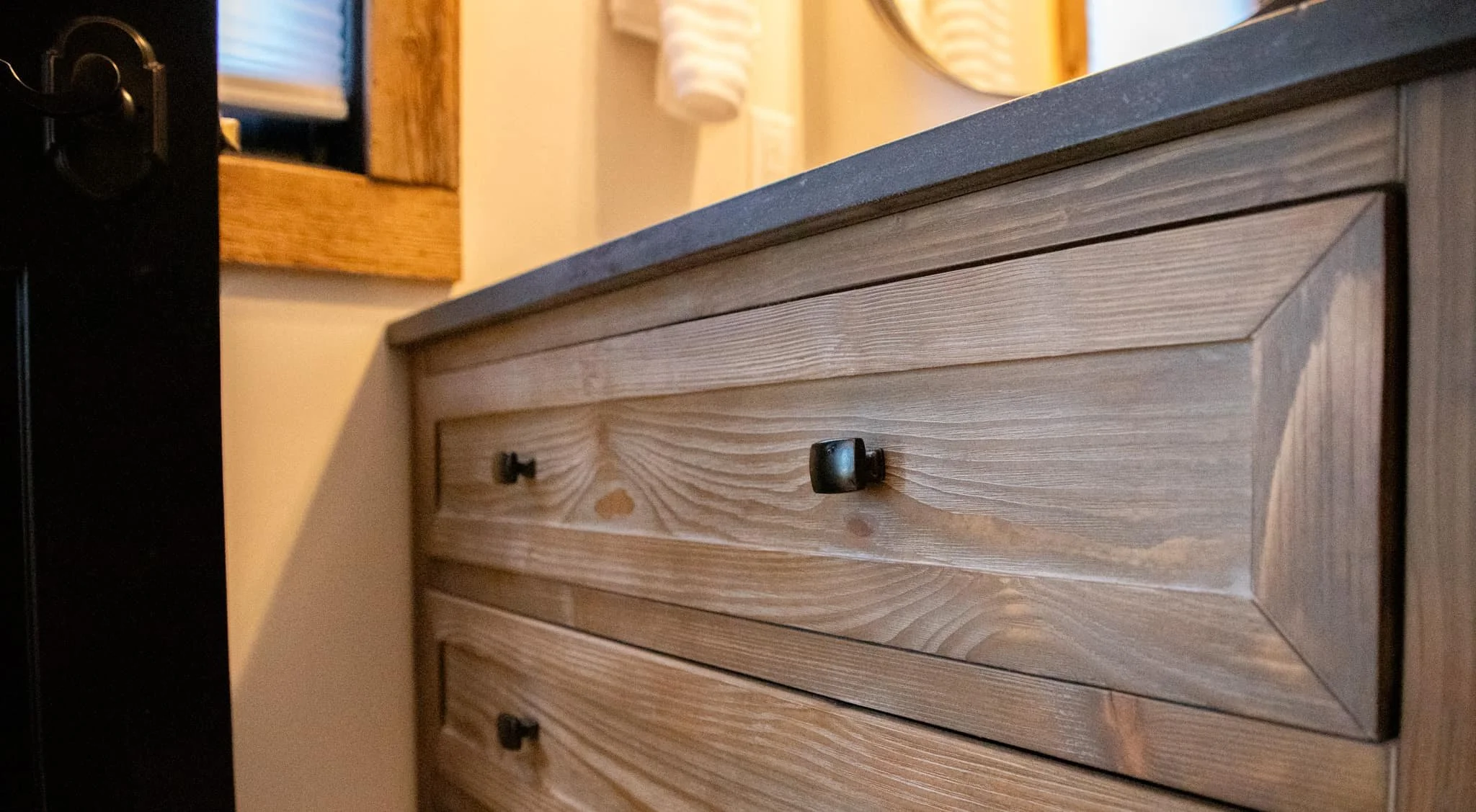 Close-up of a wooden dresser with black knobs and a top surface, in a room with a mirror and a window in the background.