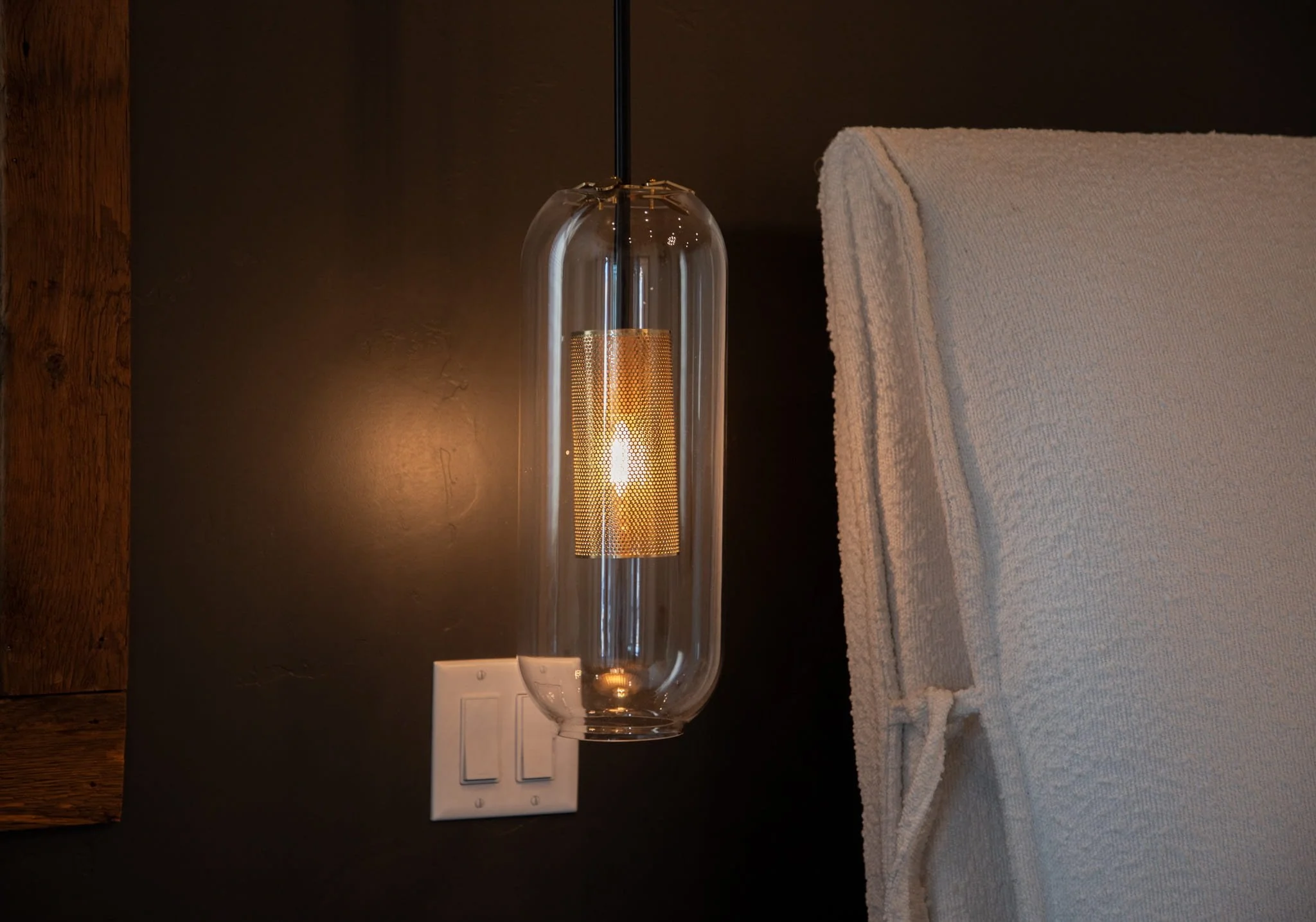 Decorative modern wall sconce with a glass enclosure and a visible Edison-style bulb, mounted on a dark wall near a bed with a beige upholstered headboard.
