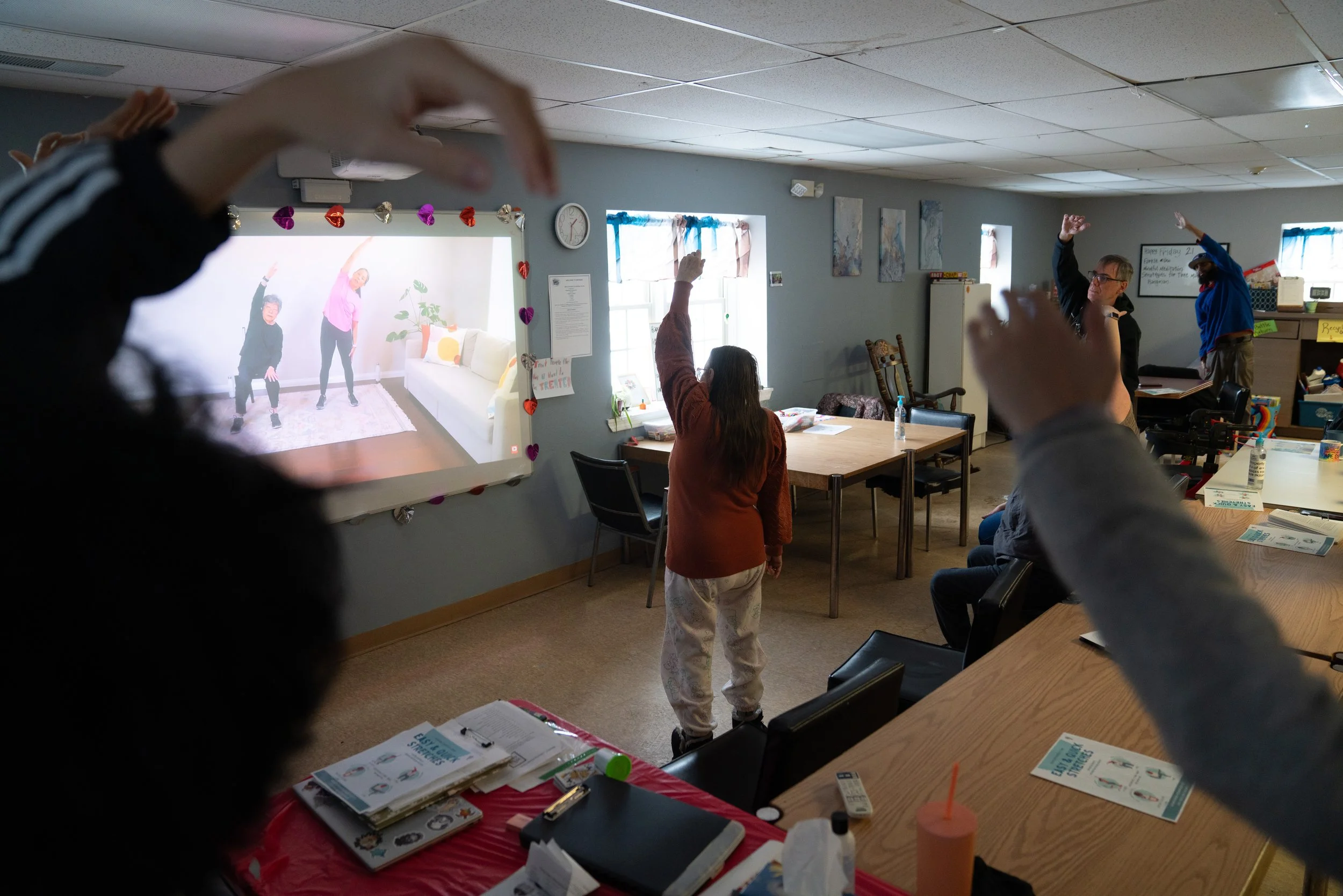 A group of people in a room participating in an exercise video displayed on a screen. Several individuals raise their arms, following the instructors on the video. The room has tables and chairs, and the screen is decorated with heart-shaped ornament