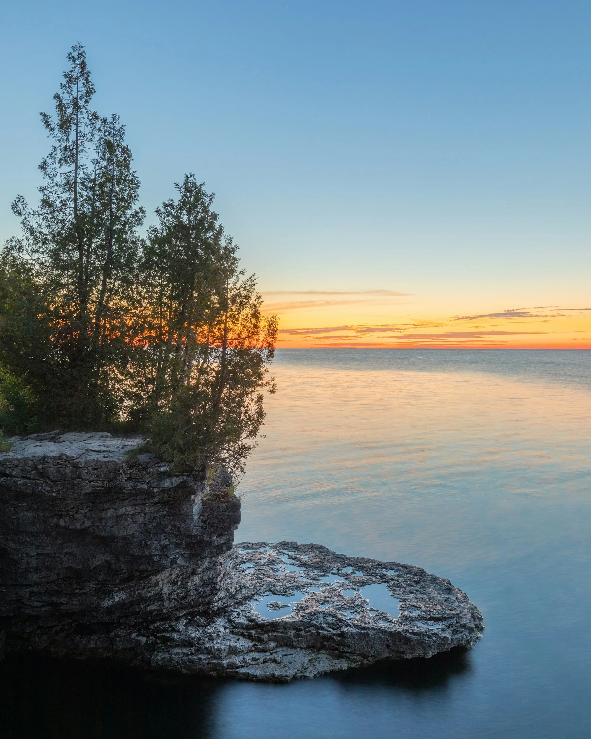 A rocky Lake Michigan shoreline with evergreen trees at sunset in Door County, Wisconsin.