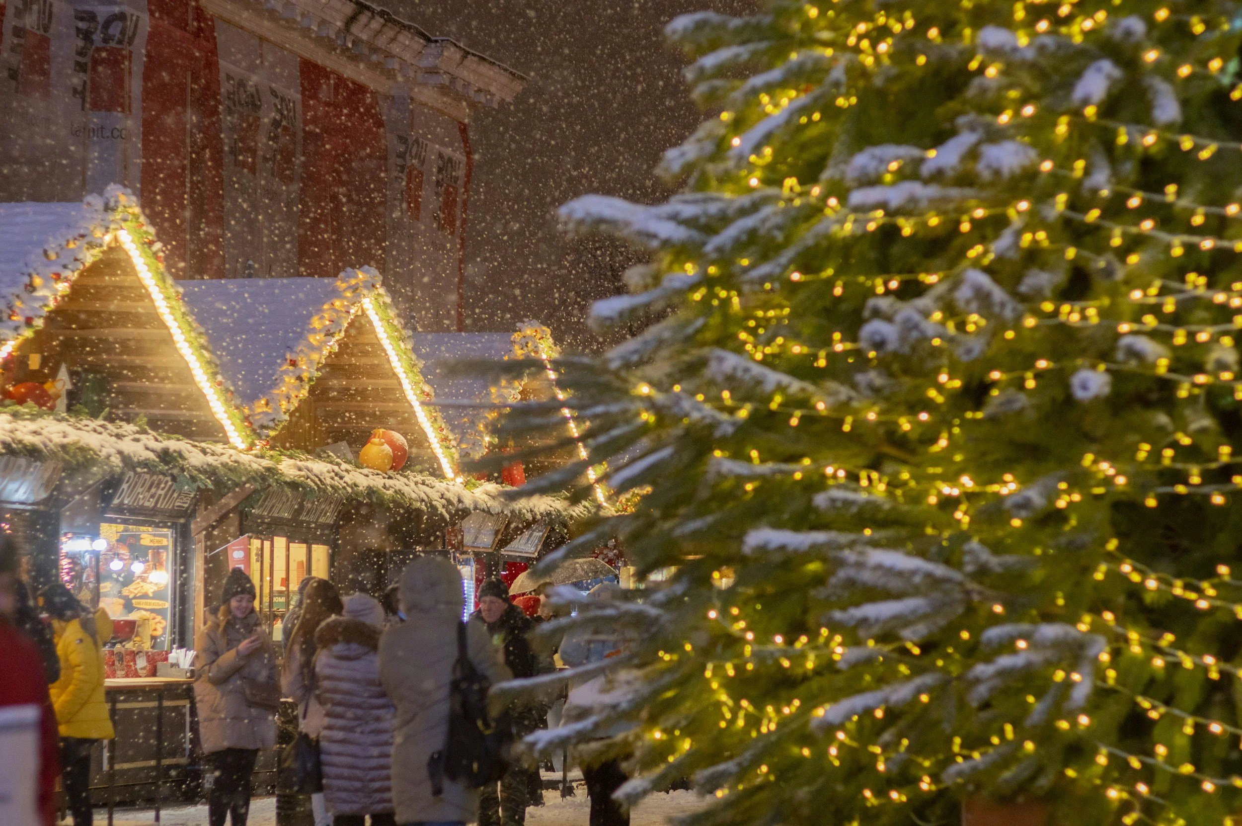 A festive outdoor Christmas market with glowing lights, evergreen trees and wooden vendor huts.
