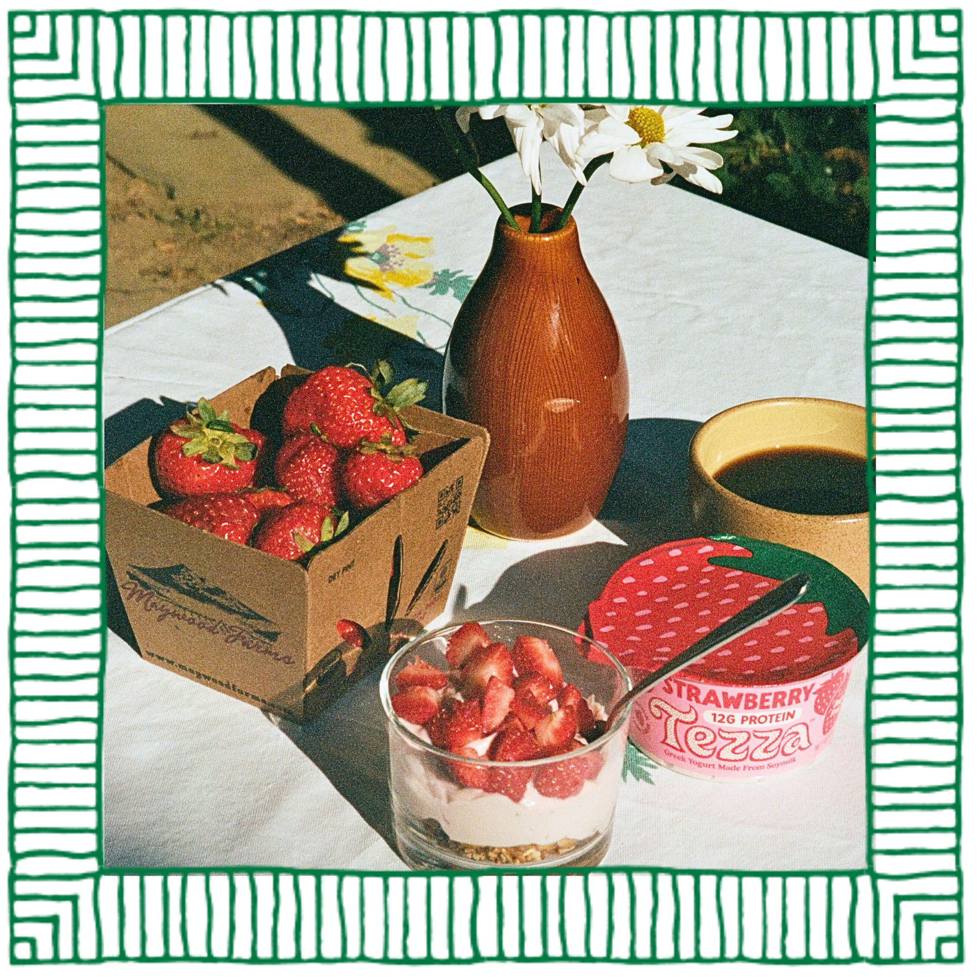 A breakfast scene with a box of fresh strawberries, a glass of yogurt with strawberries, a cup of coffee, a small yellow bowl, and a brown vase with white daisies on a table with a white tablecloth.