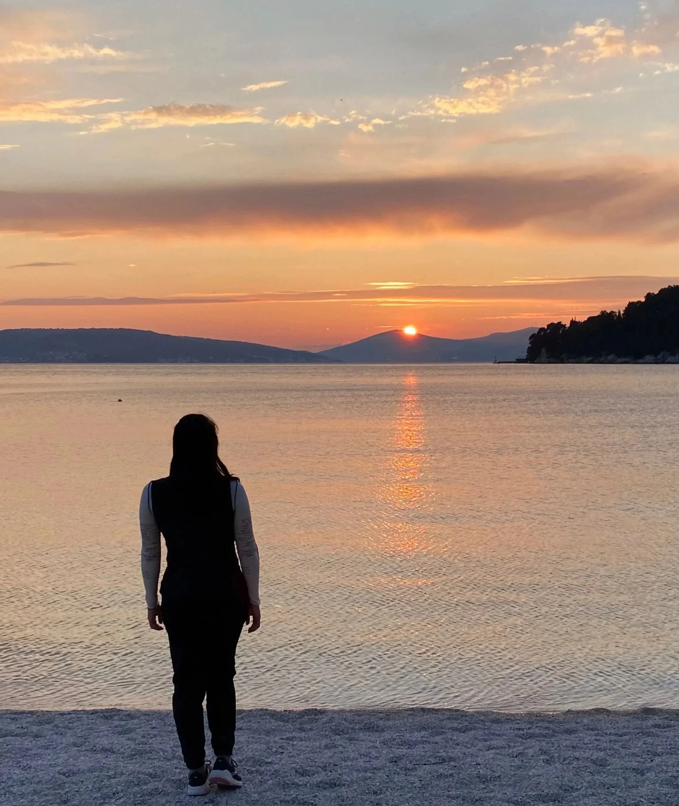 A person standing on a beach at sunset, facing a calm lake with mountains in the distance.