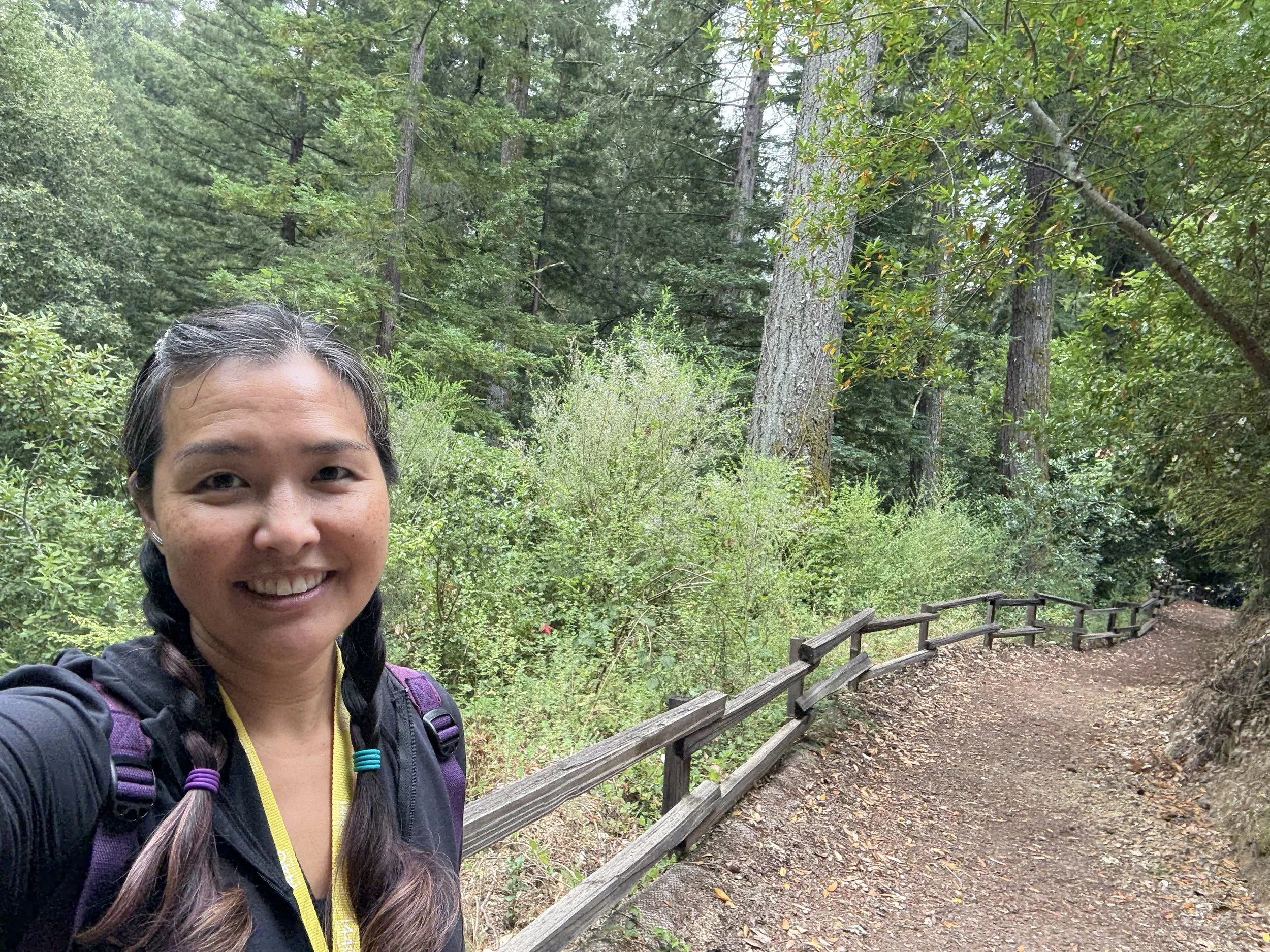A woman with braided hair taking a selfie on a forest trail with trees and greenery around.