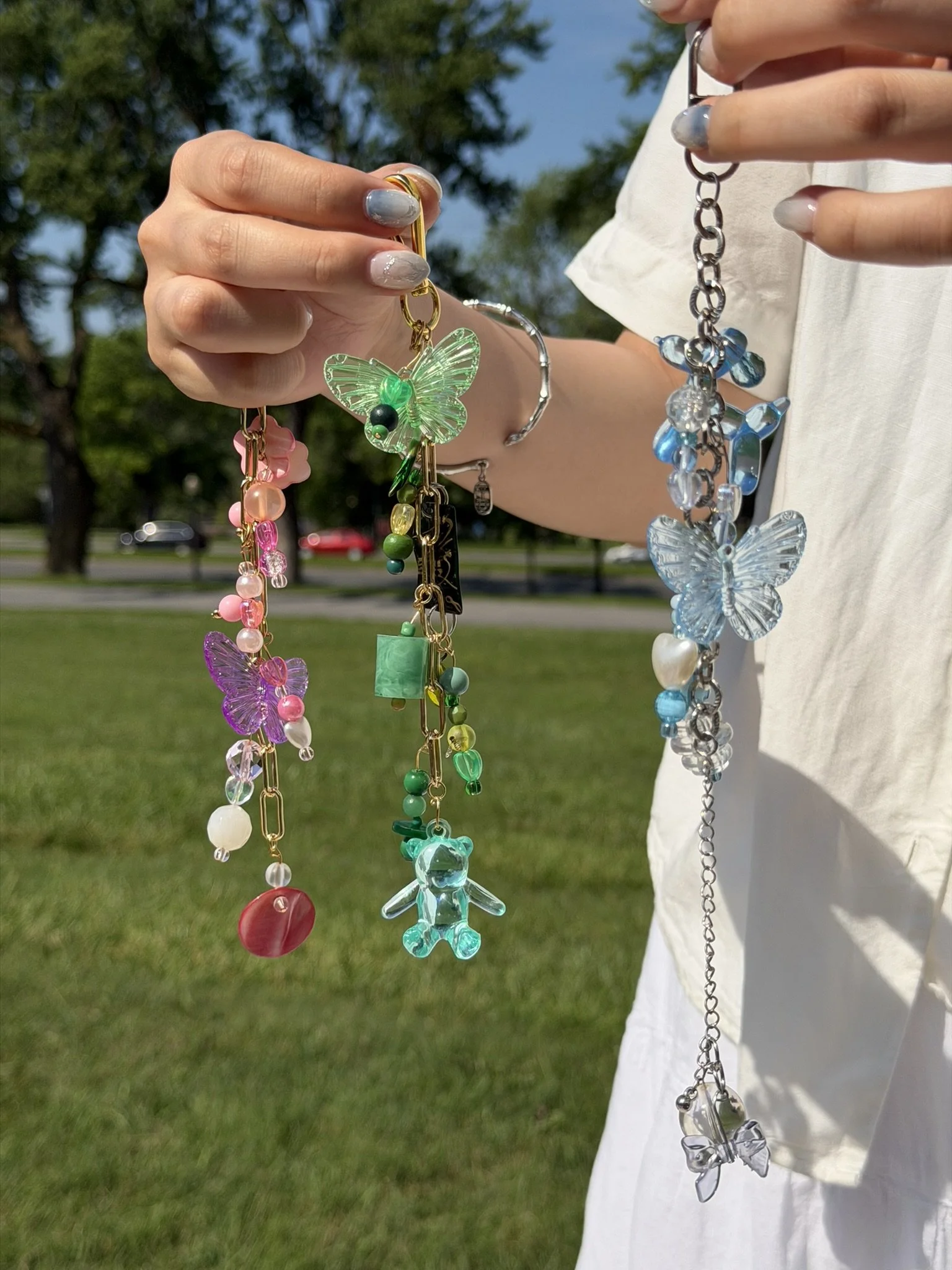 Person holding colorful beaded necklaces and bracelets outdoors, with trees and a blue sky in the background.