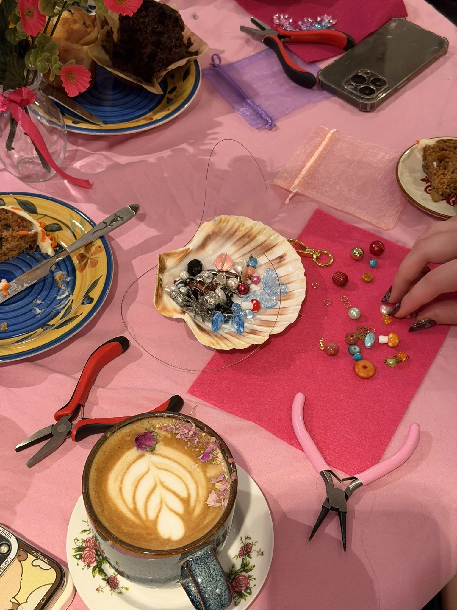 A pink table with a cup of latte with latte art, scattered colorful beads and jewelry, pliers, a smartphone, a chocolate cake, plates, and various crafting supplies.