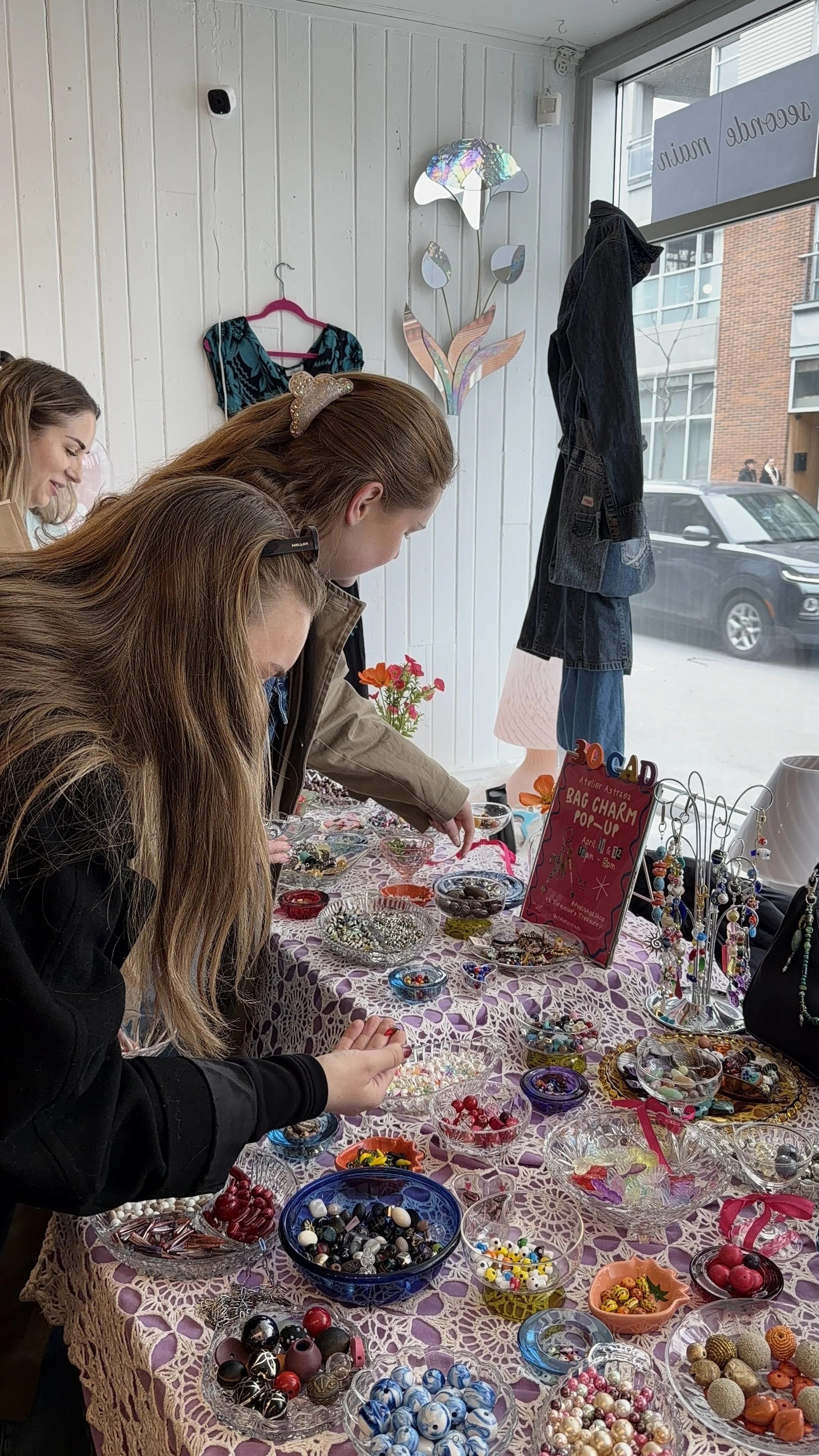 People browsing jewelry and accessories at a market stall with a table covered in a lace tablecloth, various bowls of colorful beads, buttons, and jewelry items, and a sign promoting a bag charm pop-up event.