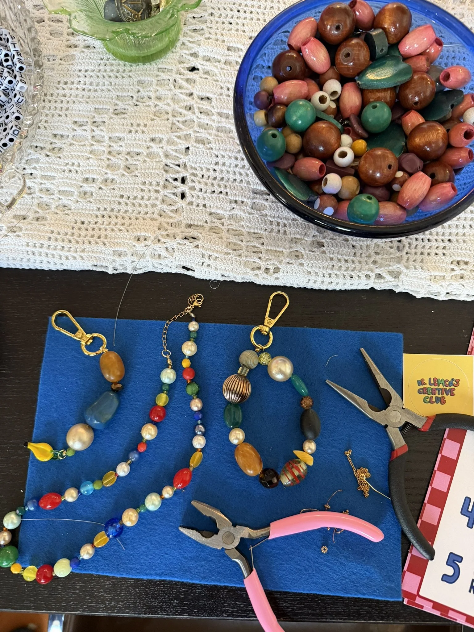 Colorful beads, jewelry-making tools, and crafting supplies on a table. A blue felt mat holds various beads and jewelry components. A pair of pliers and a pink handle tool are also visible, along with a blue card labeled 'Lemons Creative Club' and pa