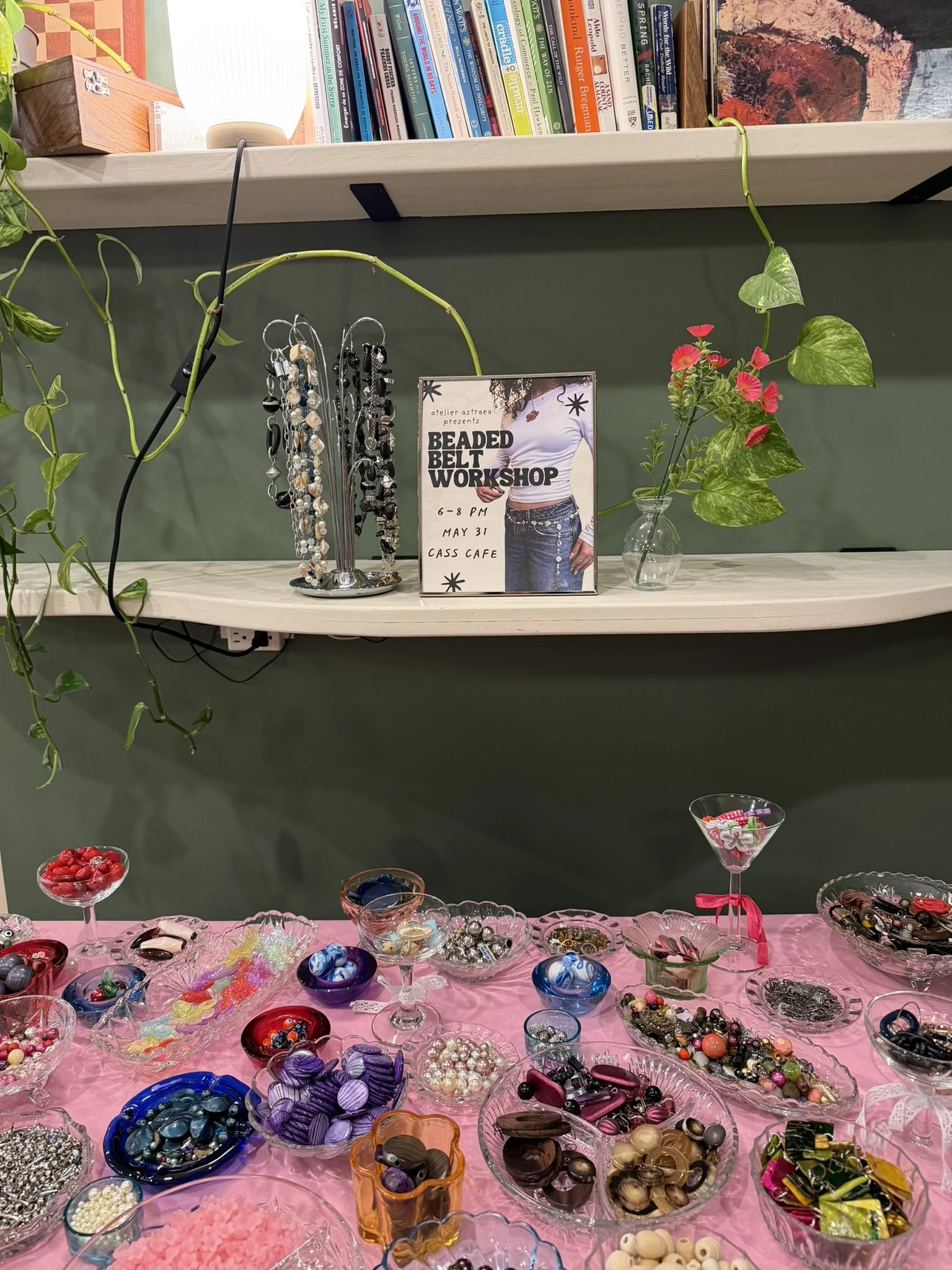 A table covered with a pink cloth displaying various beads and jewelry supplies in glass bowls. There are bowls of colorful buttons, strands of beads, and other decorative items, as well as a vase with a plant and flowers. In the background, a sign a