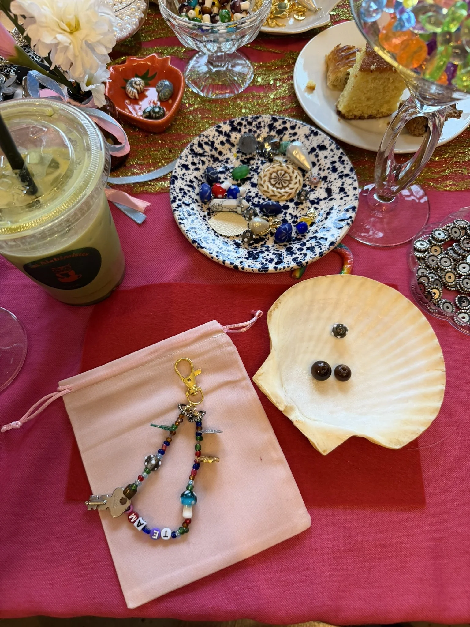 A table with a pink cloth, displaying jewelry, a pink pouch with a keychain, a shell, a drink, and various small desserts and decorations.