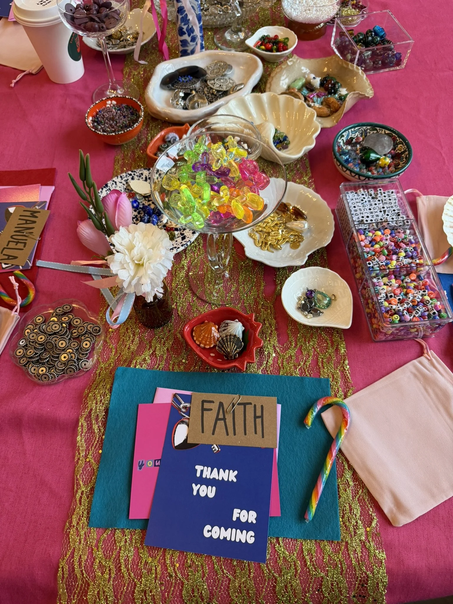 A colorful table display with glass bowls, dishes, and containers holding various candies, beads, jewelry, and decorative items, along with a bouquet of flowers, thank you cards, and a rainbow-colored candy cane.