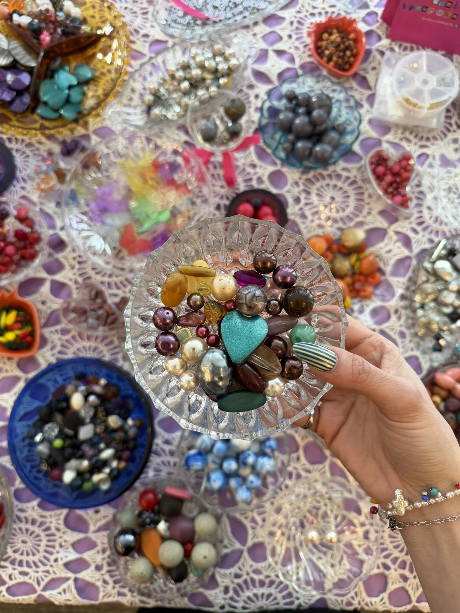 A hand holding a glass dish filled with colorful beads and jewelry charms, with various bowls of beads and jewelry pieces on a purple and white lace tablecloth in the background.