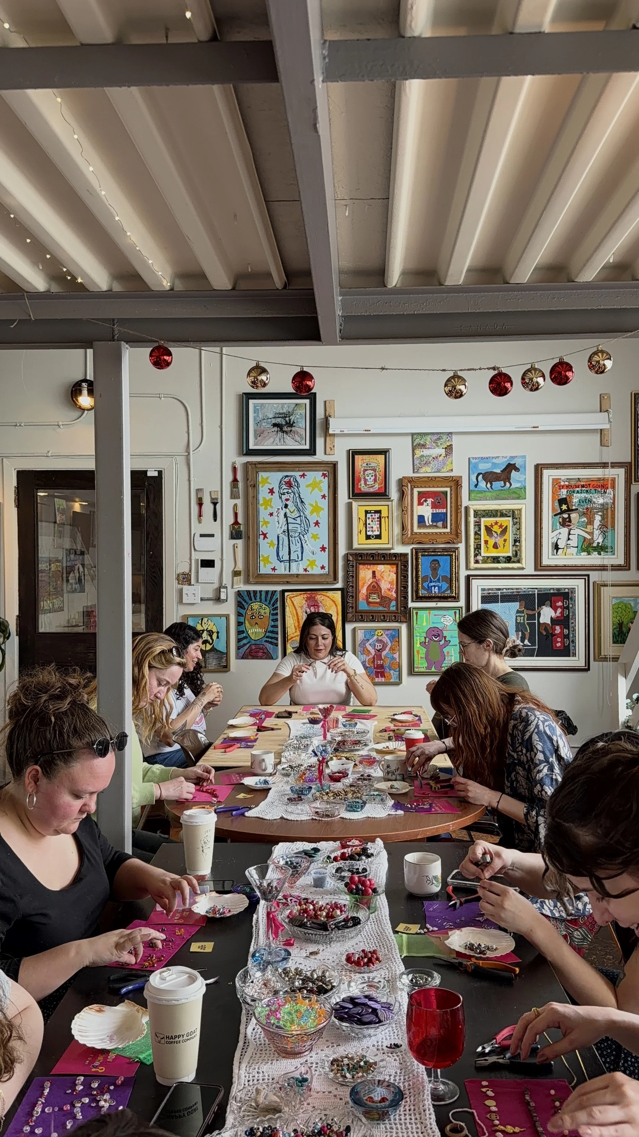 A group of women sitting around a long table decorating Christmas ornaments with beads, baubles, and other craft supplies during a festive gathering. The wall behind them features a colorful display of framed art.