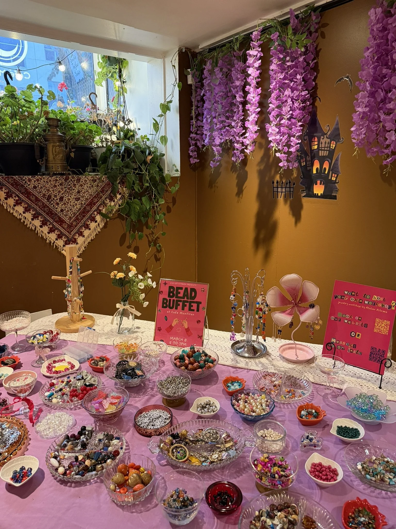 Display table filled with various beads and jewelry-making supplies, with signs advertising a bead buffet and a jewelry workshop, decorated with purple hanging flowers and a Halloween-themed paper decoration on the wall.