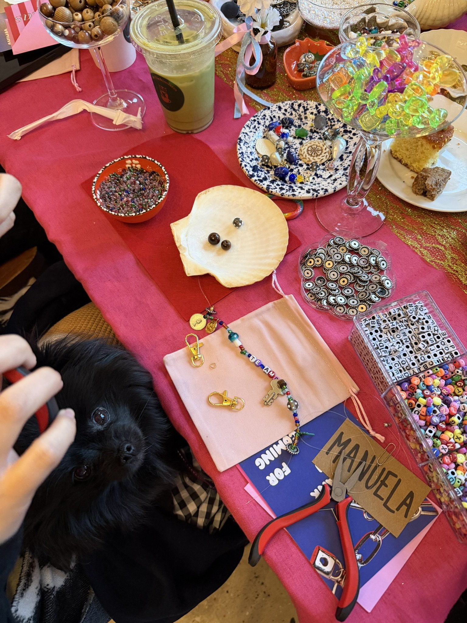 A table with various craft supplies, jewelry, candies, and a black puppy with shiny eyes being held by a person. The tablecloth is red, and the background has decorative items and a pair of red scissors.