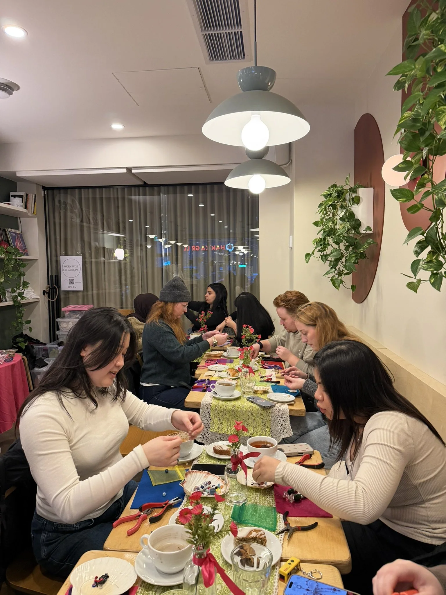 People sitting at a long table in a decorated room, engaged in a craft activity, with tea cups, plates, and crafting supplies on the table, and plants and wall decorations in the background.