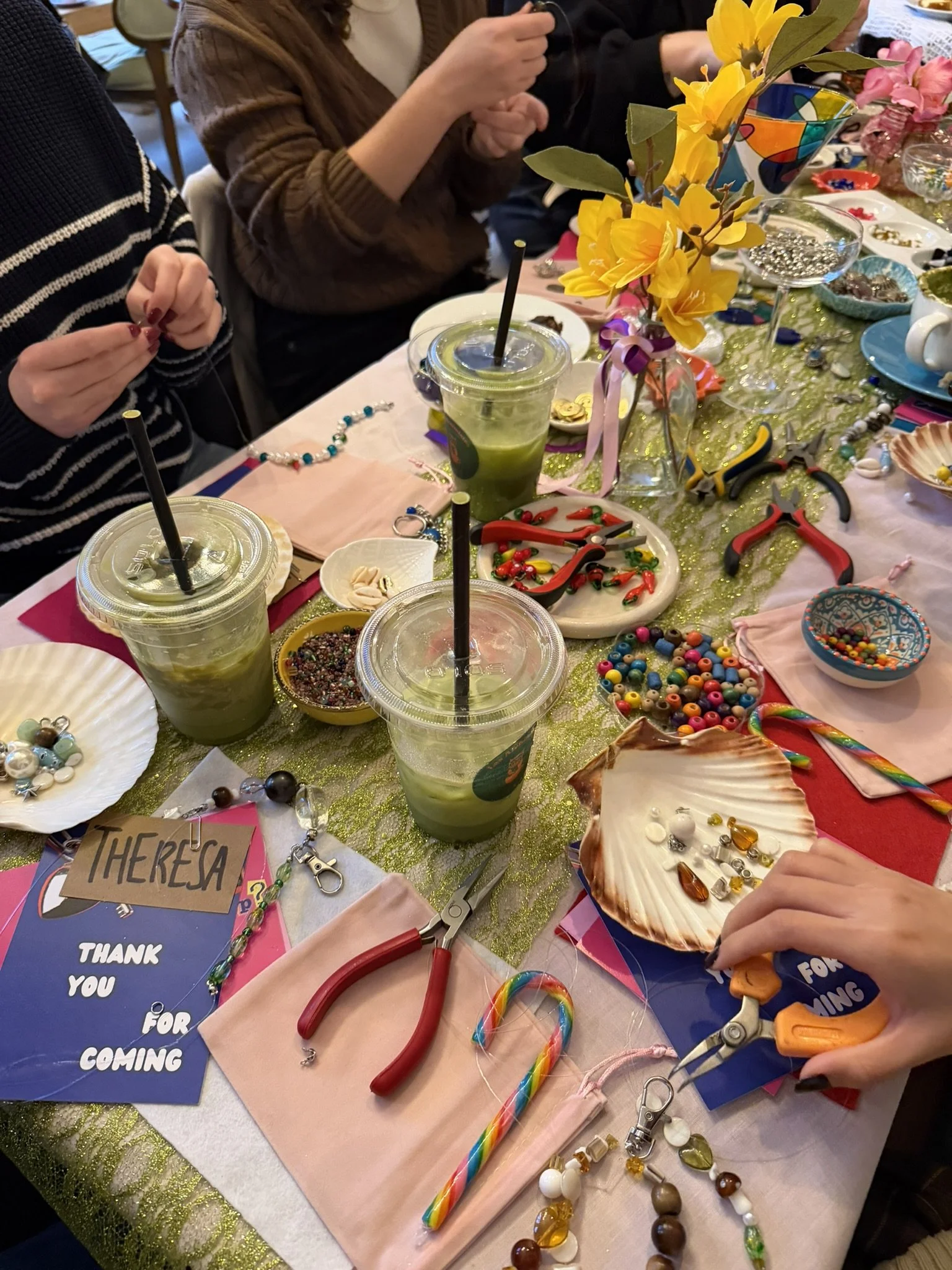 A decorated table with various crafting supplies, including colorful beads, pliers, and elastic cords, along with drinks and flowers, at a crafting or jewelry-making event.