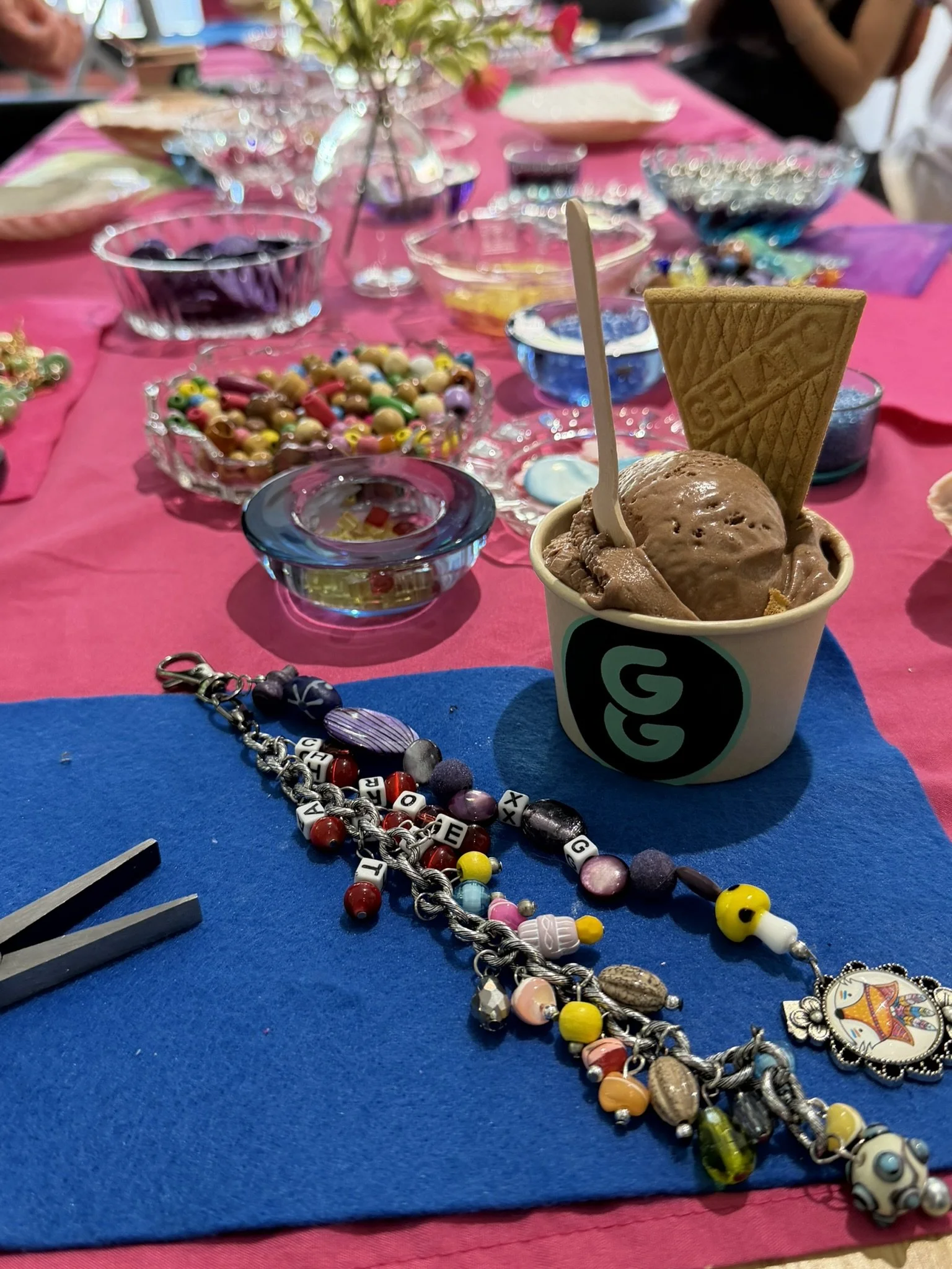 Ice cream with cone and spoon on a colorful table at a craft or jewelry event, with beads and candy decorations nearby.