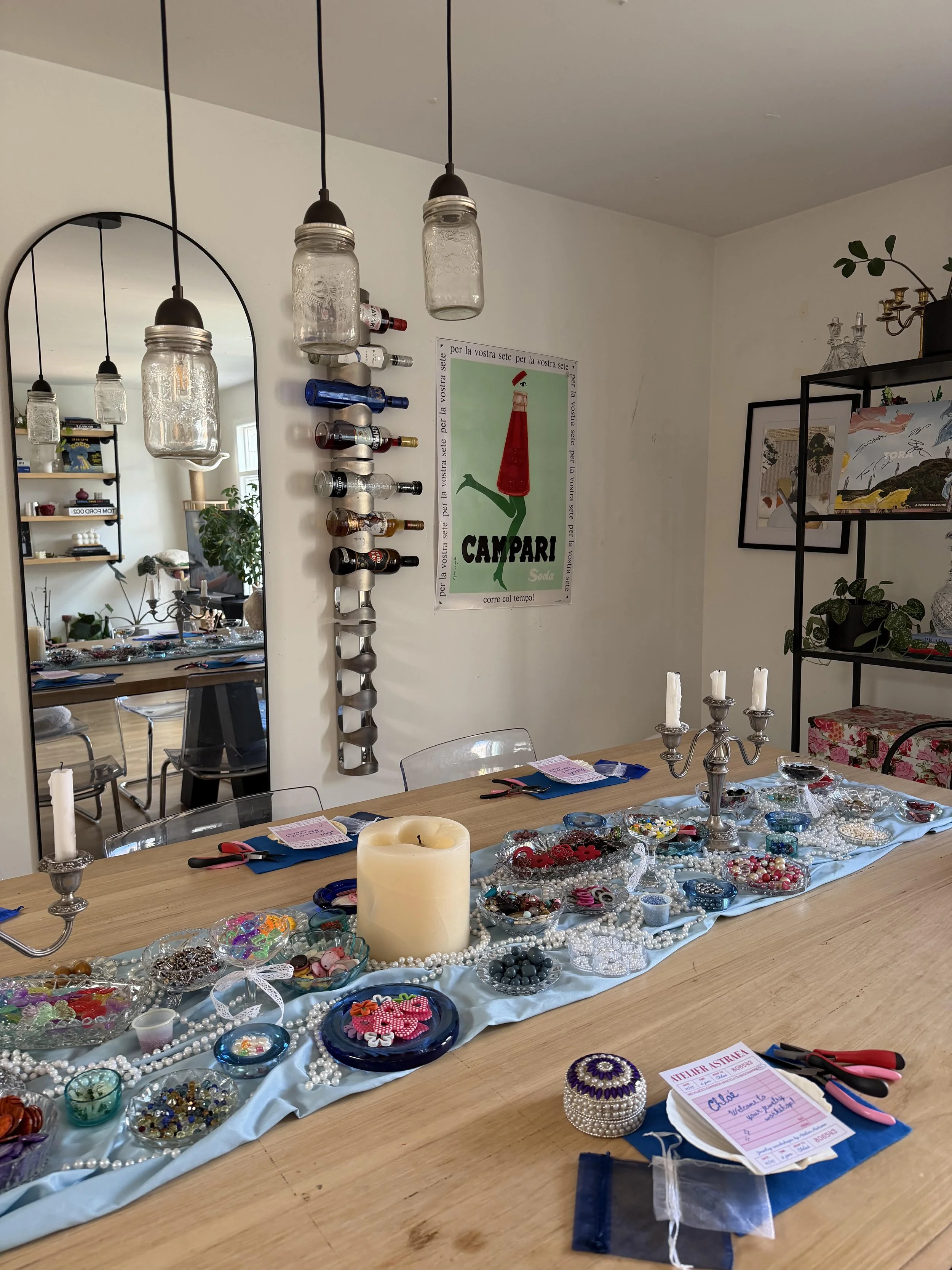 A dining table covered with various jewelry pieces, including necklaces, bracelets, and beads, with a large candle in the center and a note card nearby. In the background, there is a wall with a poster, a wine rack, and decorative items in a living room.
