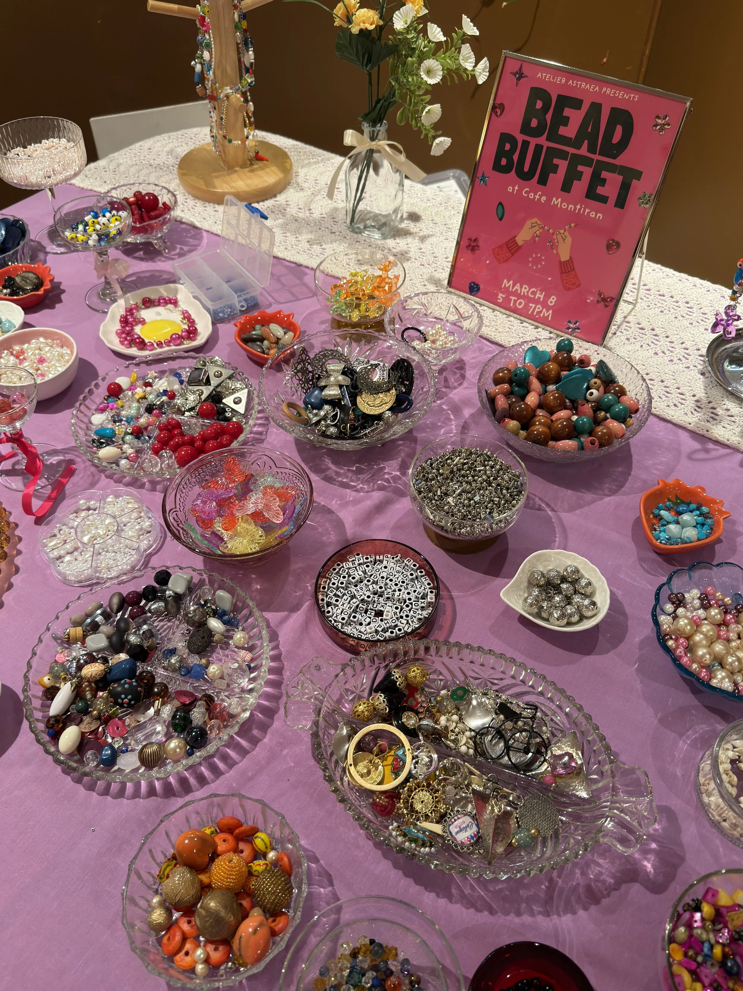 Display table with various bowls and containers filled with colorful beads, buttons, and jewelry components, with a pink event sign reading 'Bead Buffet' at Cafe Montiran.