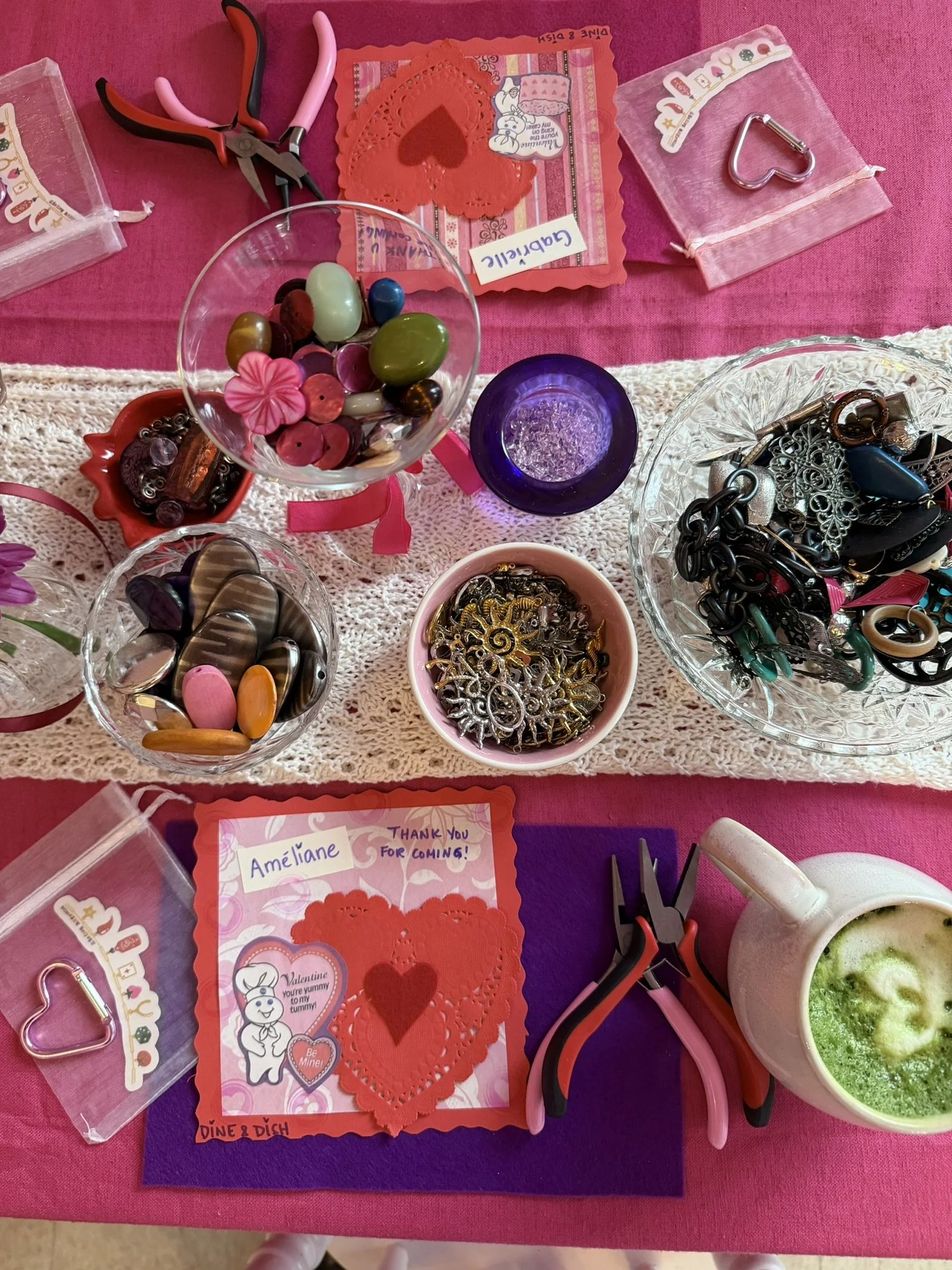 A table displays Valentine's Day craft supplies, including heart-shaped doilies, stickers, containers of buttons and jewelry parts, scissors, and a cup of green-colored beverage or snack, all on a pink and purple tablecloth.