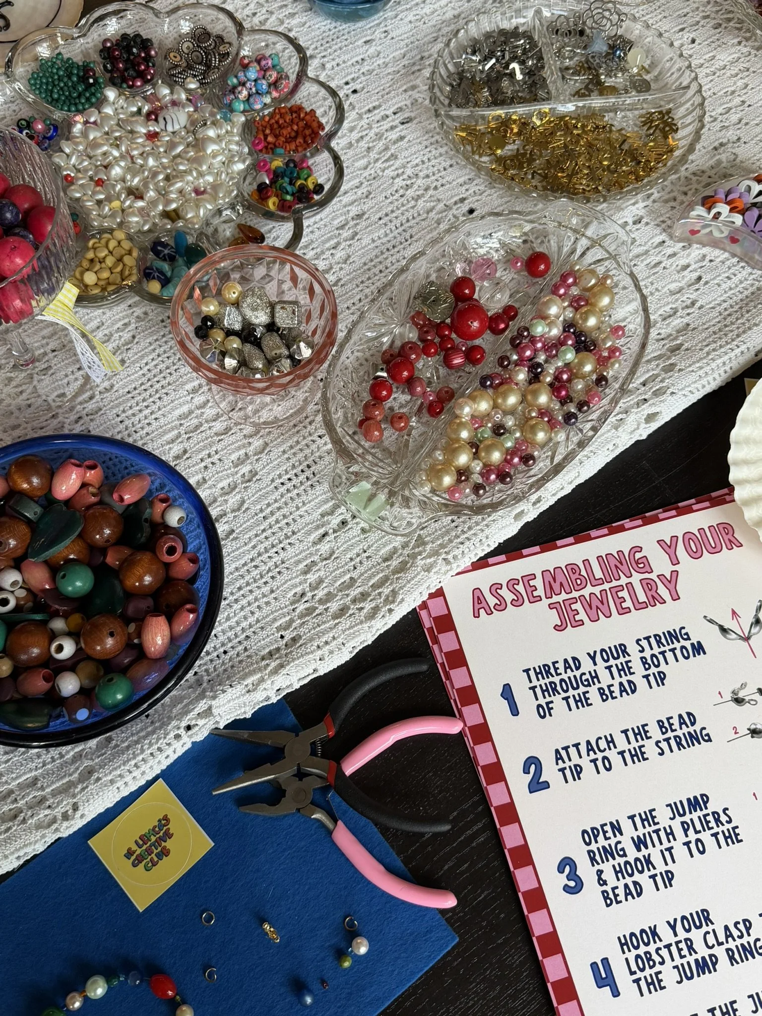 Variety of jewelry-making beads in glass dishes, jewelry pliers, and instructions for assembling jewelry on a table.