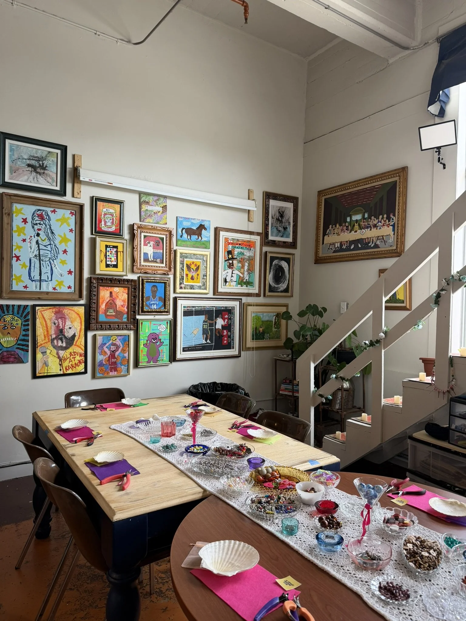 A dining area with a table set for a craft or jewelry-making activity. The table has colorful napkins, scissors, and various beads and jewelry supplies. The room features a wall decorated with framed artwork and a staircase with candles. The space is