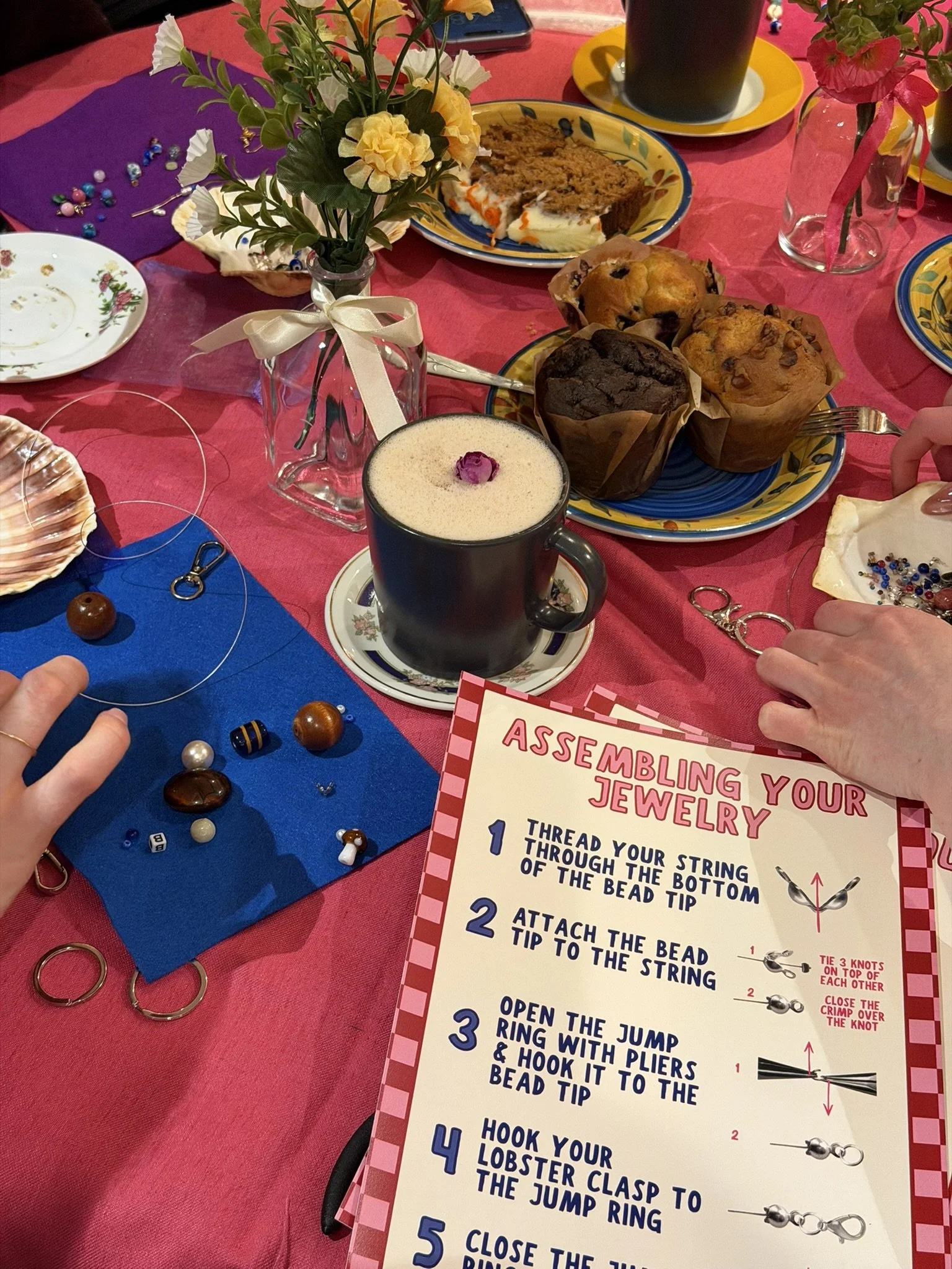 A table set for a gathering with various desserts, a flower arrangement, and a jewelry-making activity. The tablecloth is pink, and there are colorful plates, cups, and decorations.