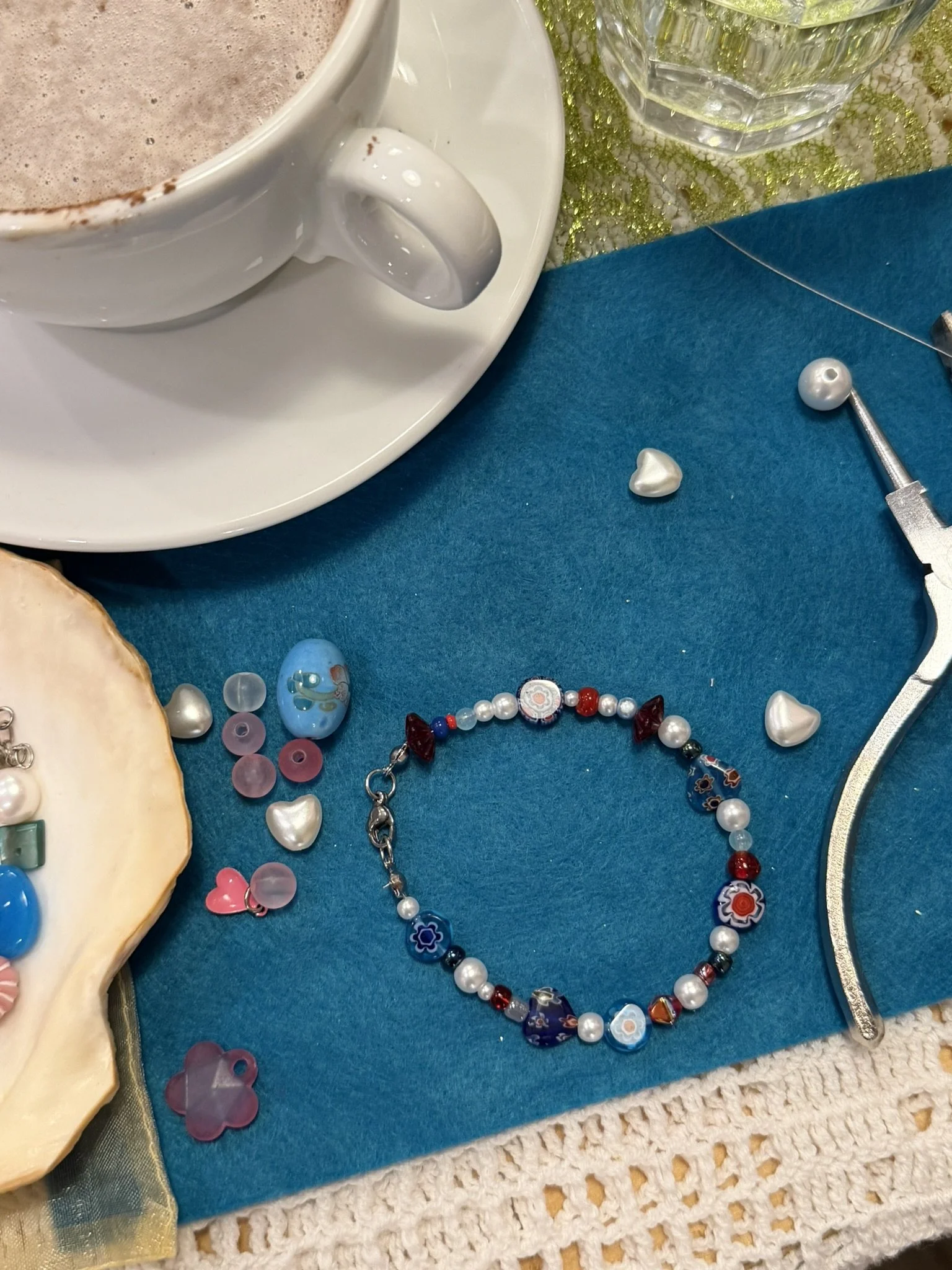 Close-up of jewelry, a white cup of hot chocolate, a glass of water, and decorative items on a blue surface with a gold-trimmed cloth underneath.