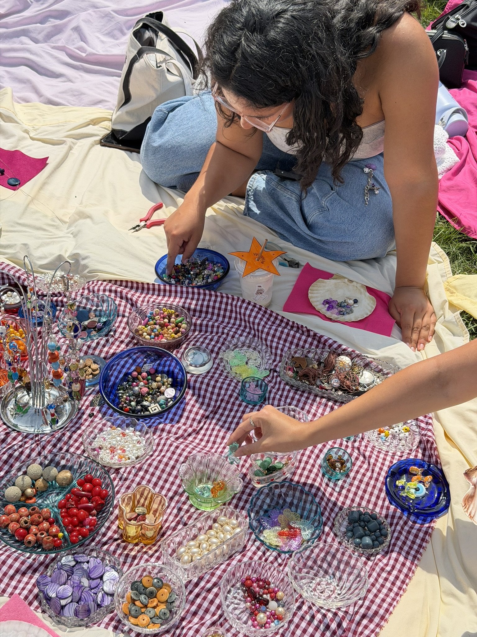 A person with curly hair browsing buttons at an outdoor craft fair table with various bowls of colorful buttons and craft supplies.