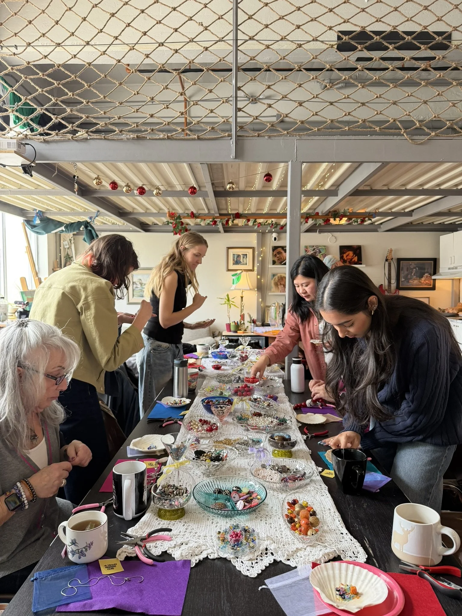 Group of people gathered around a long table making crafts or jewelry. The table is covered with beads, buttons, and craft supplies, and the room is decorated with holiday ornaments and string lights.