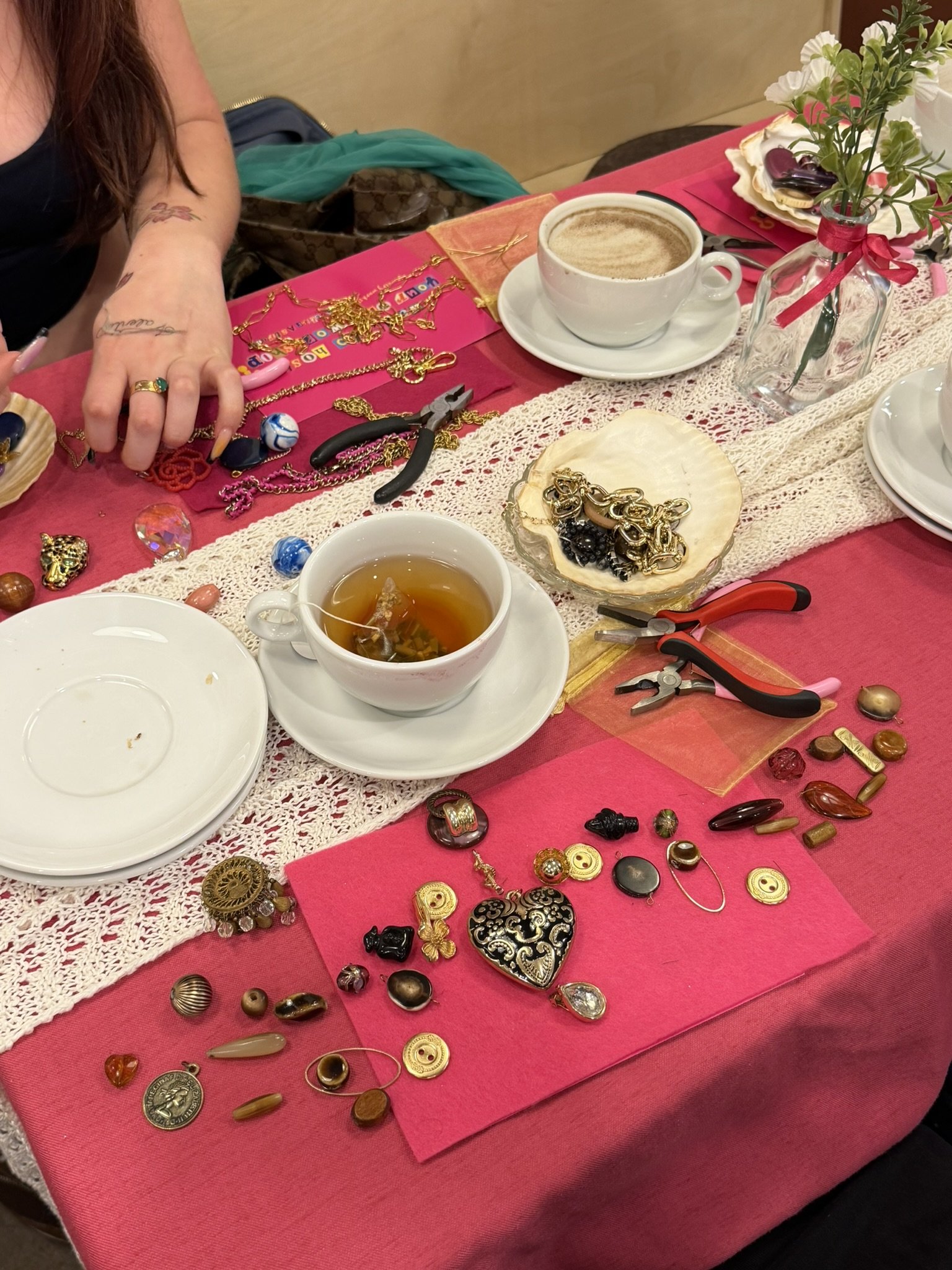 A table with various jewelry pieces including rings, necklaces, pins, and brooches. There are two cups of tea or coffee, a small vase with flowers, a pair of scissors, and a few plates. The table is covered with a pink tablecloth and a lace runner.