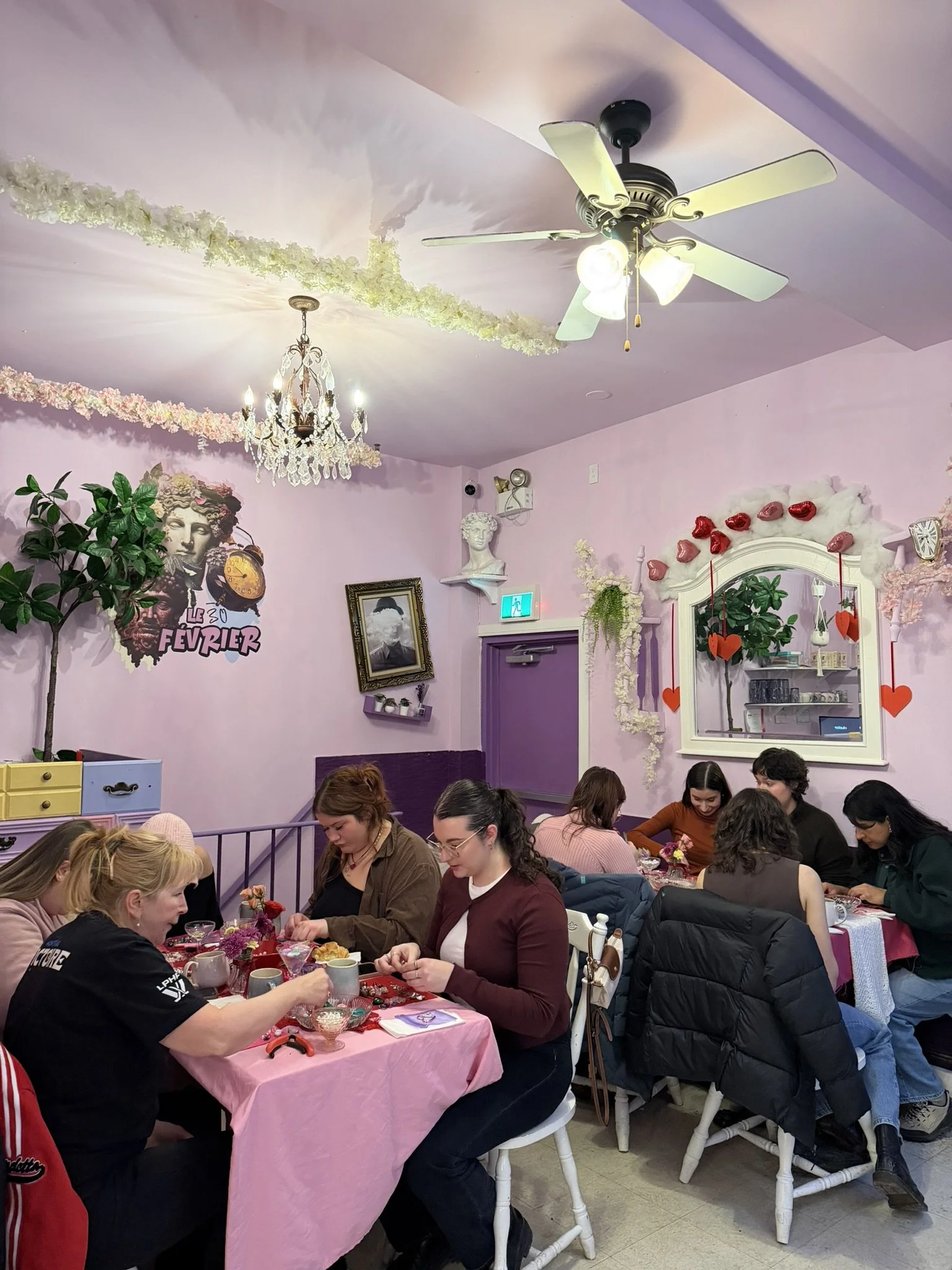 A group of people sitting around a long pink table in a decorated room, engaging in a craft activity. The room has pastel pink walls, a chandelier, floral garlands on the ceiling, and wall decorations including a statue bust, framed picture, and a la