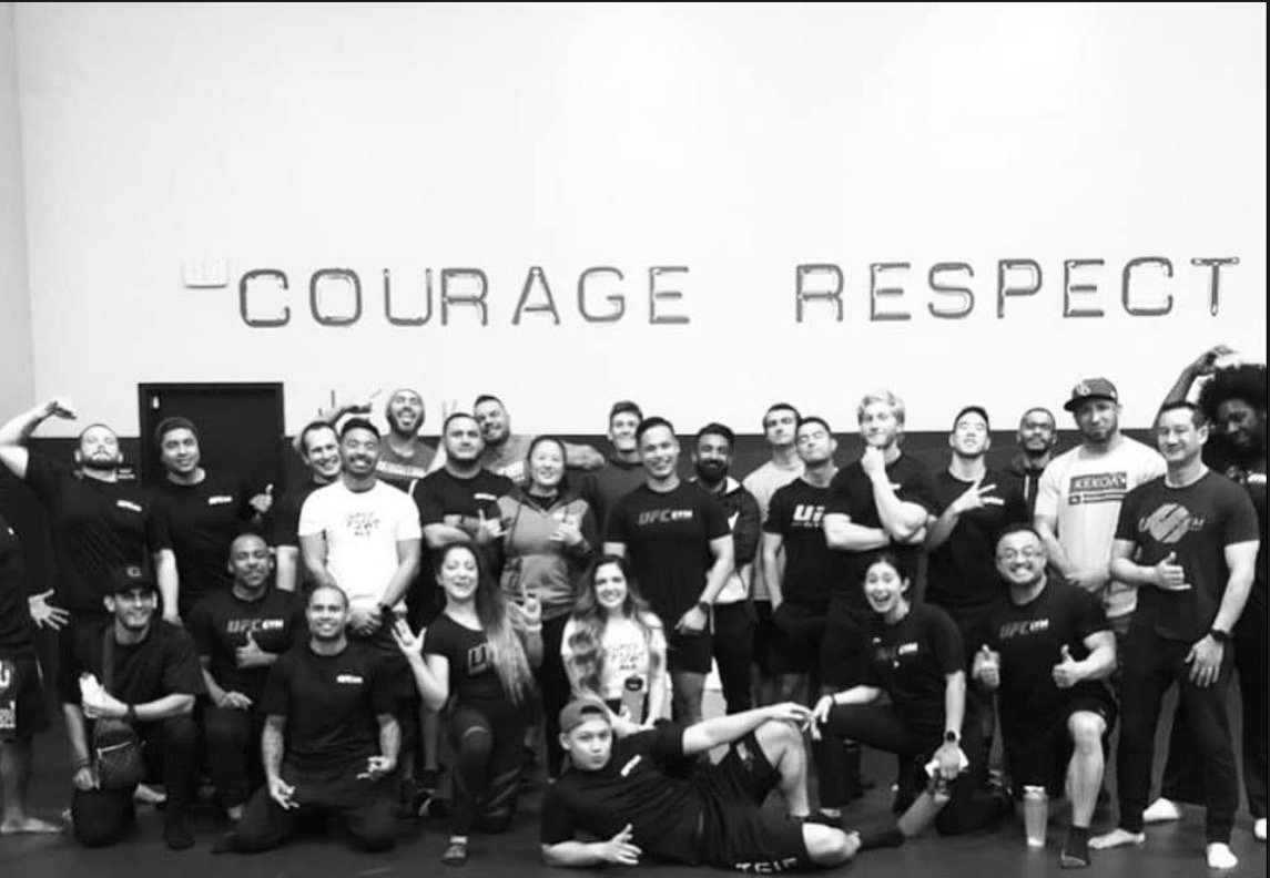 Group of people posing in front of a screen displaying the words 'COURAGE RESPECT' with some individuals flexing and smiling.