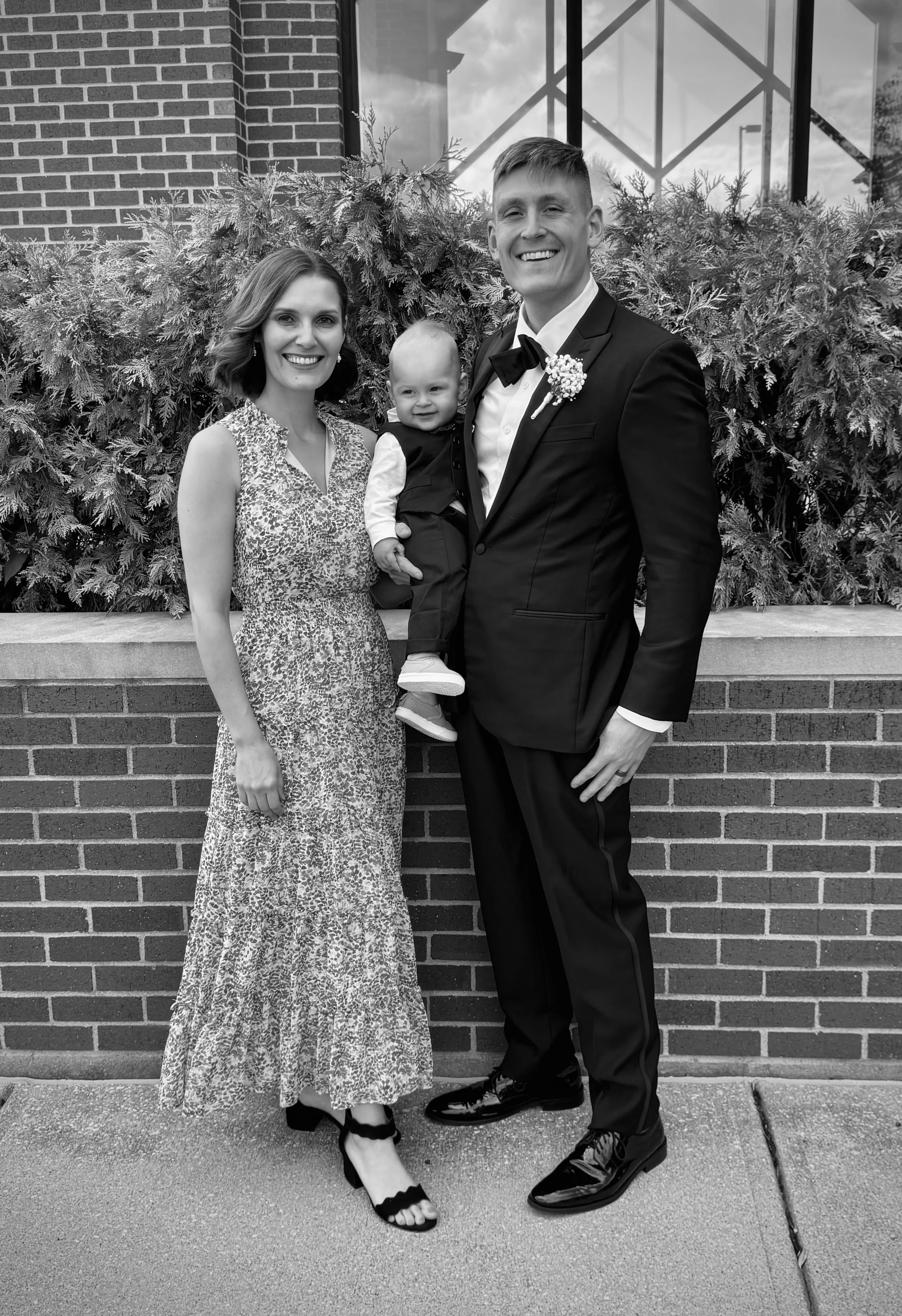 A family of three standing in front of a brick wall with bushes behind them. The woman on the left has shoulder-length hair, is wearing a patterned dress, and has high-heeled shoes. The man on the right is dressed in a tuxedo with a bow tie and a flower boutonniere. He is holding a smiling young child, who is dressed in a vest, shirt, and pants, with sneakers. The scene appears to be at a formal event, possibly a wedding.