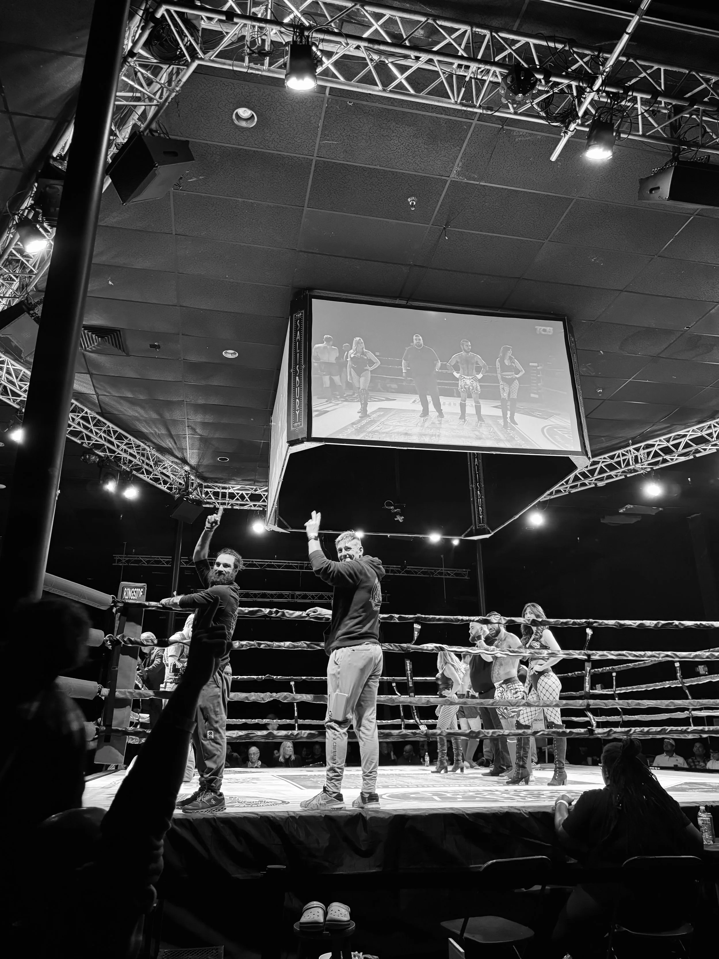 A black and white photo of a boxing ring with several people inside, some holding up hands, and a large screen hanging above showing the fighters in the ring. Audience members and officials are visible around the ring.