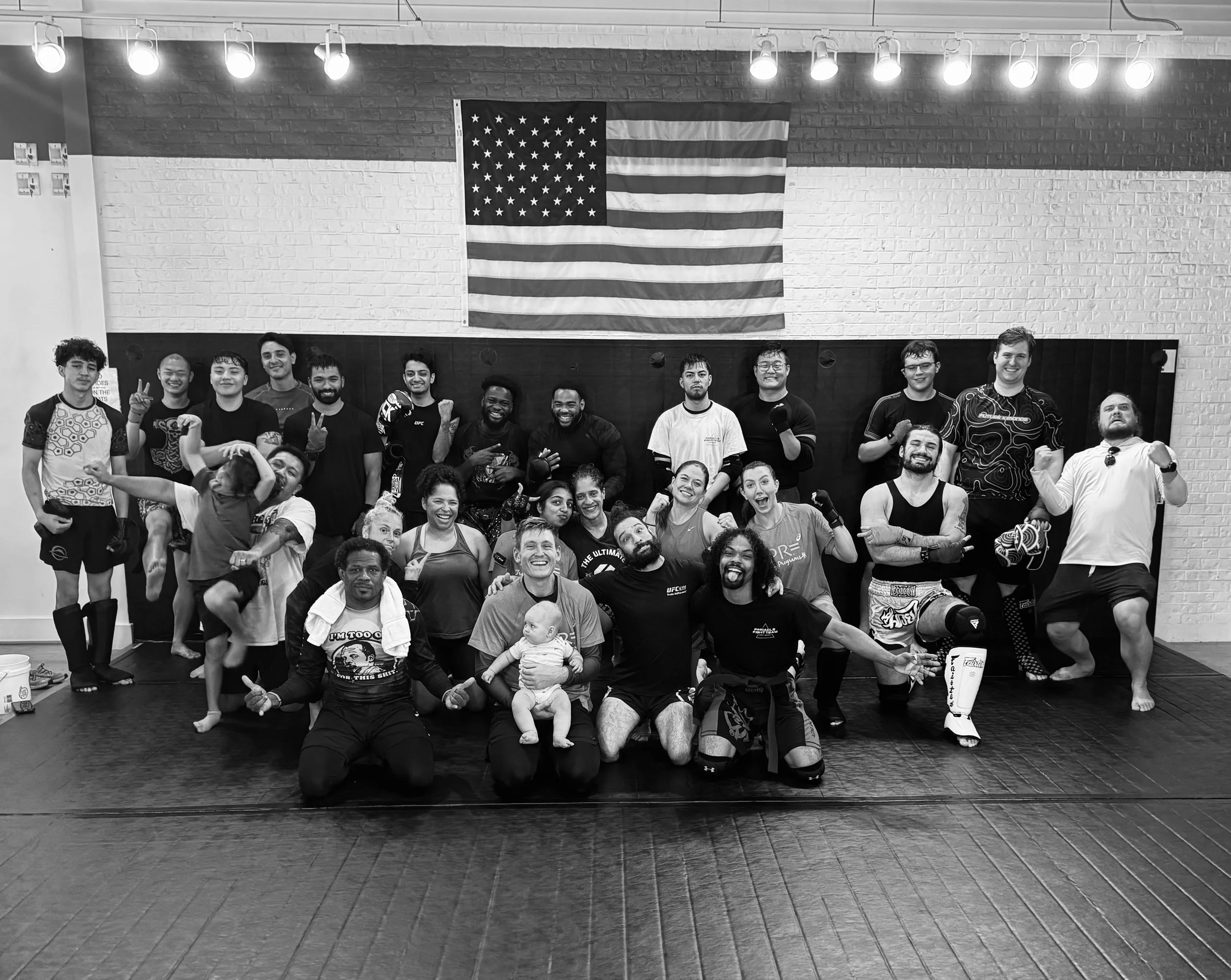 Group of people in a gym, some wearing mixed martial arts apparel, in front of an American flag on a brick wall, some smiling and holding up peace signs.