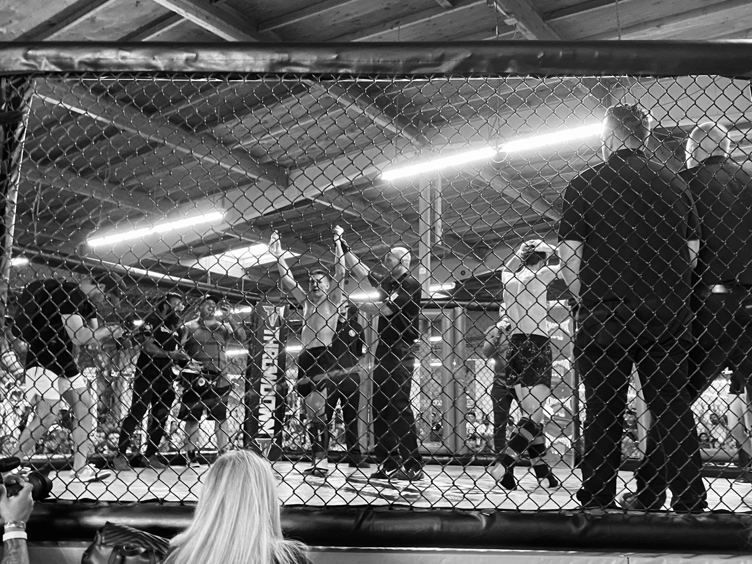A mixed martial arts fight inside a cage with chain-link fencing, with fighters, referees, and spectators visible in the background.