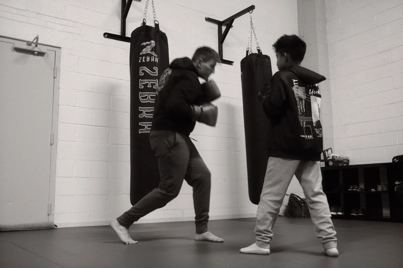 Two young men practicing boxing in a gym. One is throwing a punch while the other is holding boxing pads. There are punching bags hanging from the ceiling, a door on the left, and a shelf with shoes on the right.